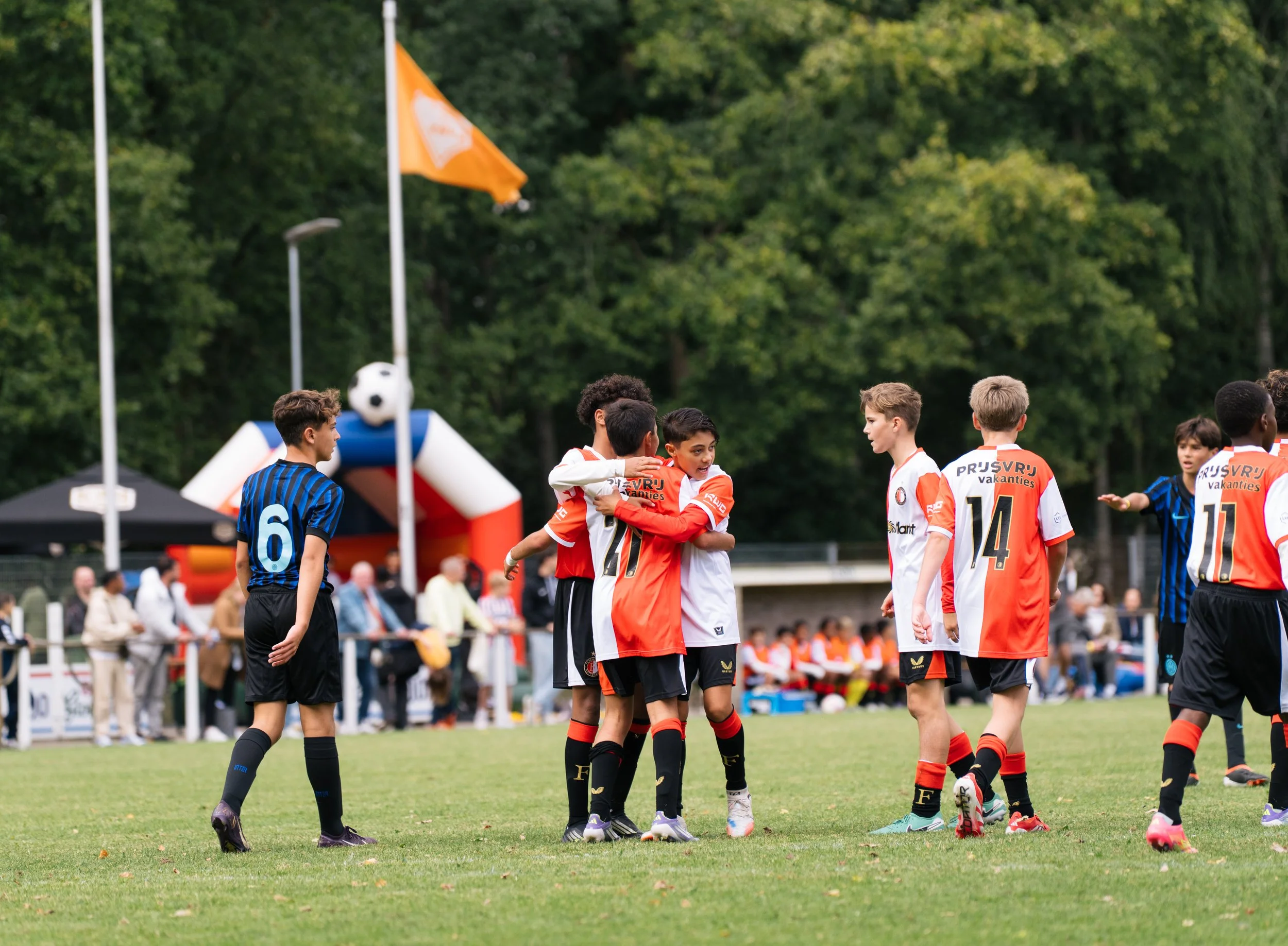 Young Feyenoordplayers in red and white jerseys embrace and talk on a soccer field during a game. Other players and spectators are visible in the background with a large inflatable building and flags. During the Virgil's Legacy Trophy tournament in T