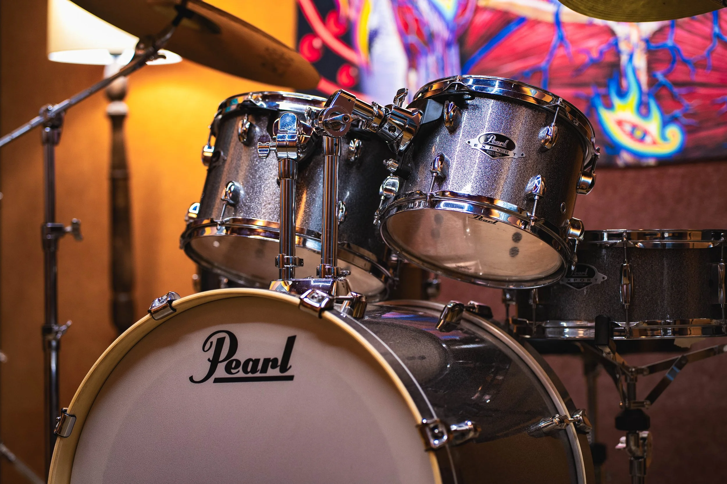 A close-up of a Pearl brand drum set with a cymbal and drumsticks on a stand in a music room, with colorful artwork on the wall in the background.