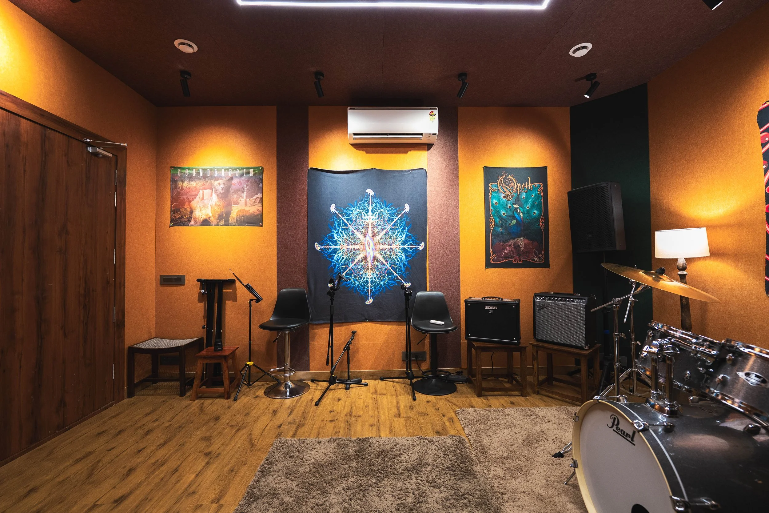 An indoor music rehearsal space with orange walls adorned with band posters, including Opeth and Soundgarden. The room features a drum set, microphone stands, two chairs, an amplifier, and a speaker. A small lamp provides additional lighting, and the
