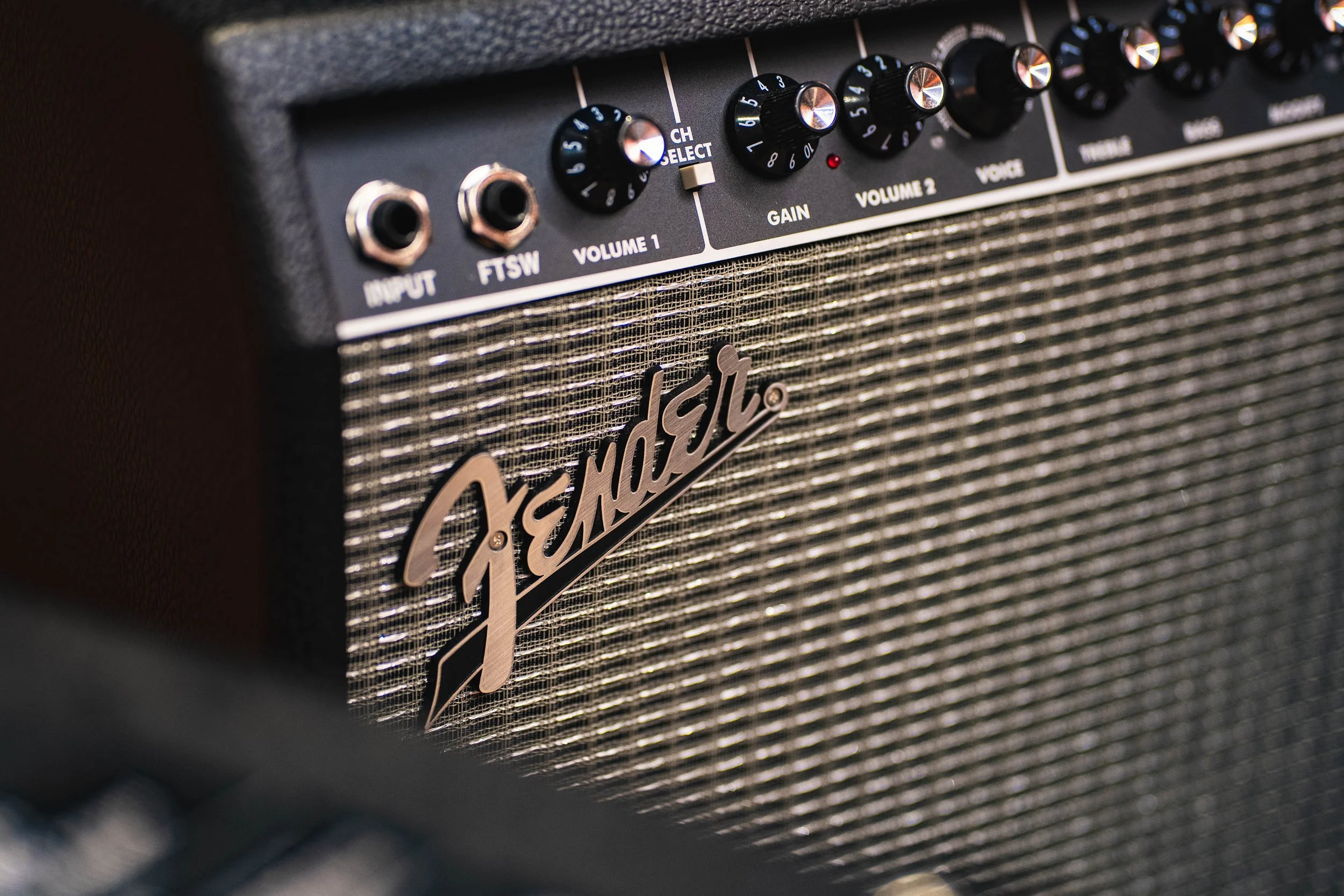 Close-up of a Fender guitar amplifier showing knobs and controls on the top panel, with the Fender logo on the front grille.
