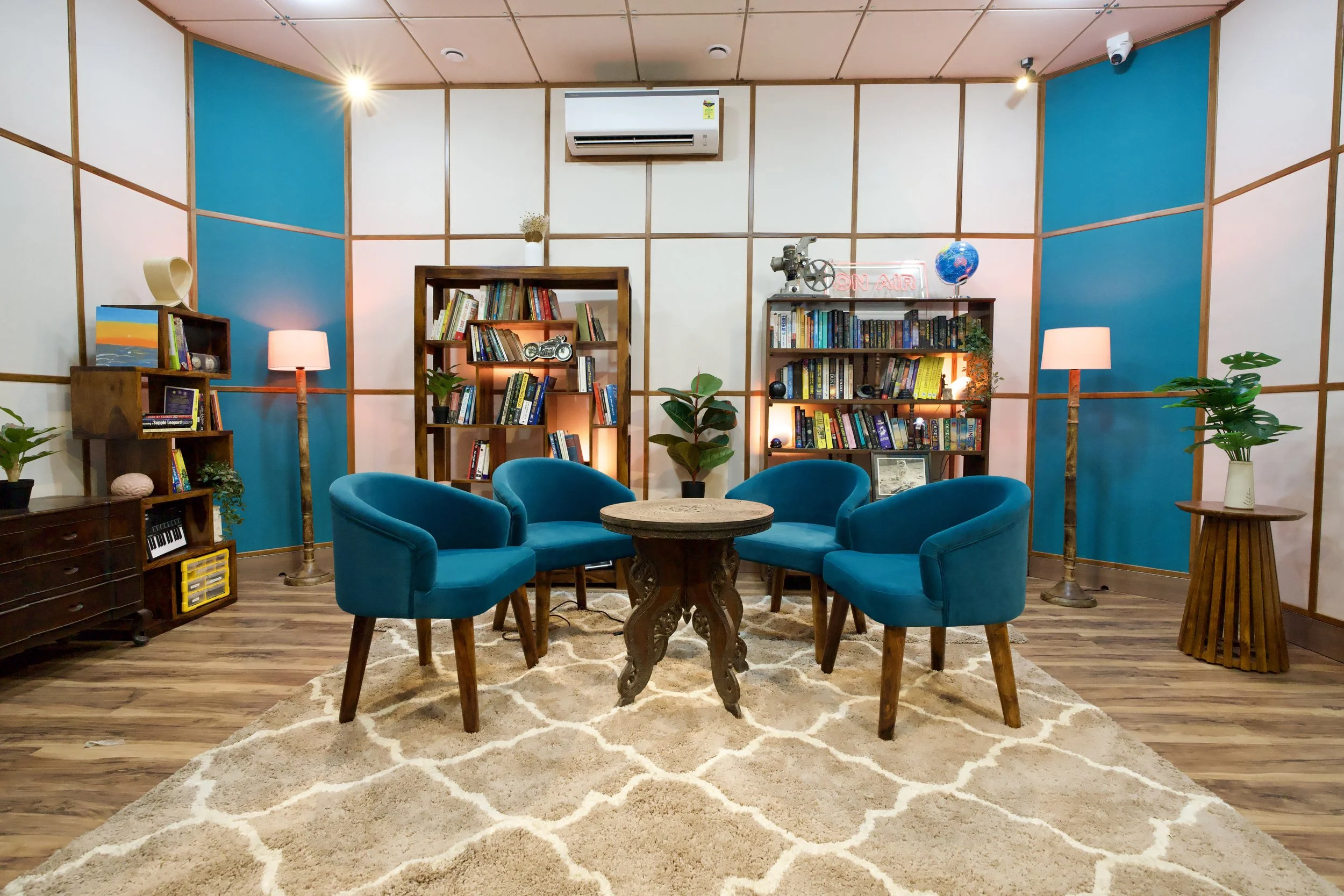 A cozy, modern library or lounge room with blue accent walls, wooden shelves filled with books, a round wooden table with four blue upholstered chairs, potted plants, lamps, and decorative items on shelves, a beige patterned rug, and an air condition