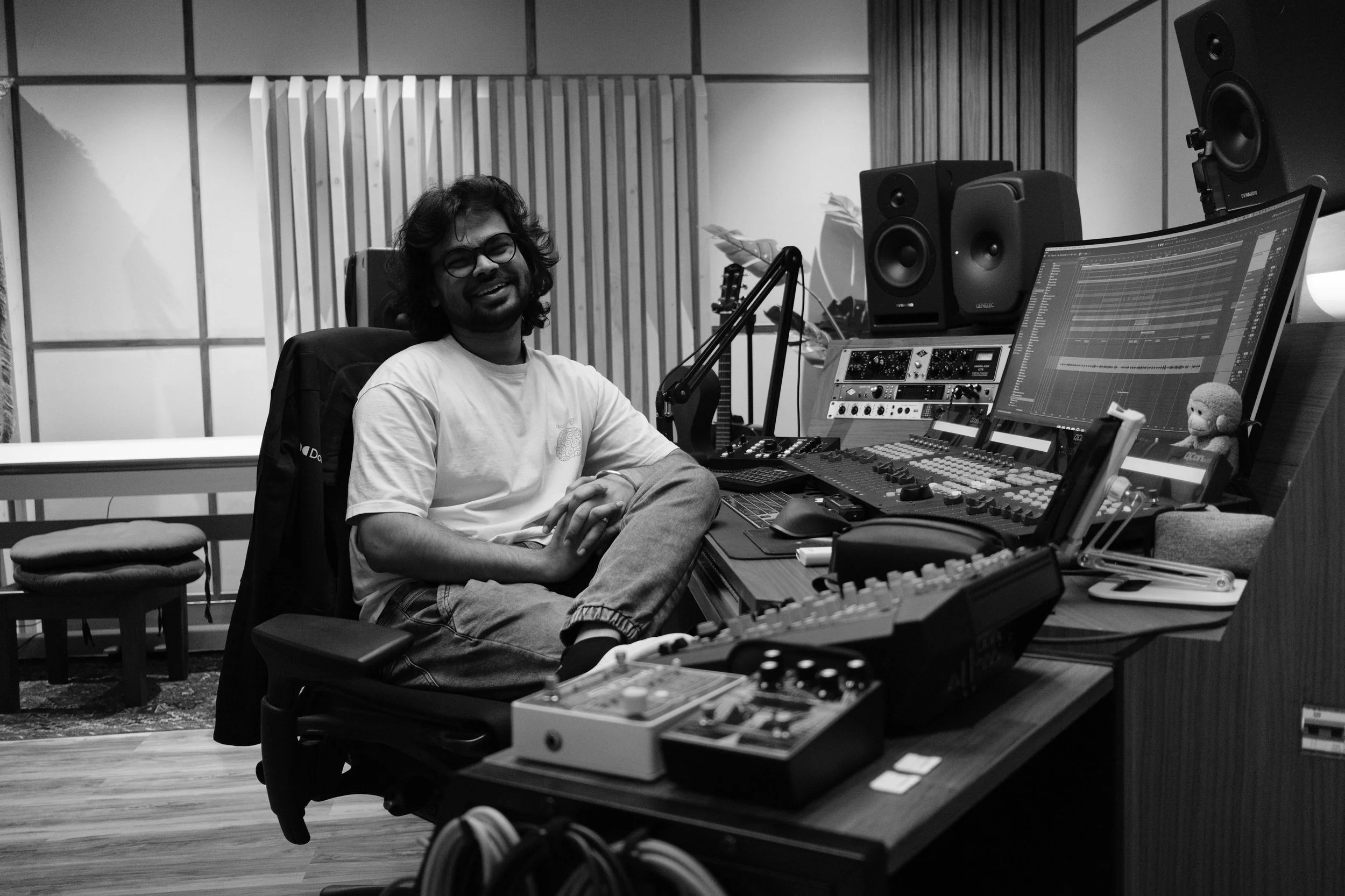 A man with glasses and long hair sitting in a recording studio surrounded by professional audio equipment, including speakers, a mixing console, and a computer monitor.