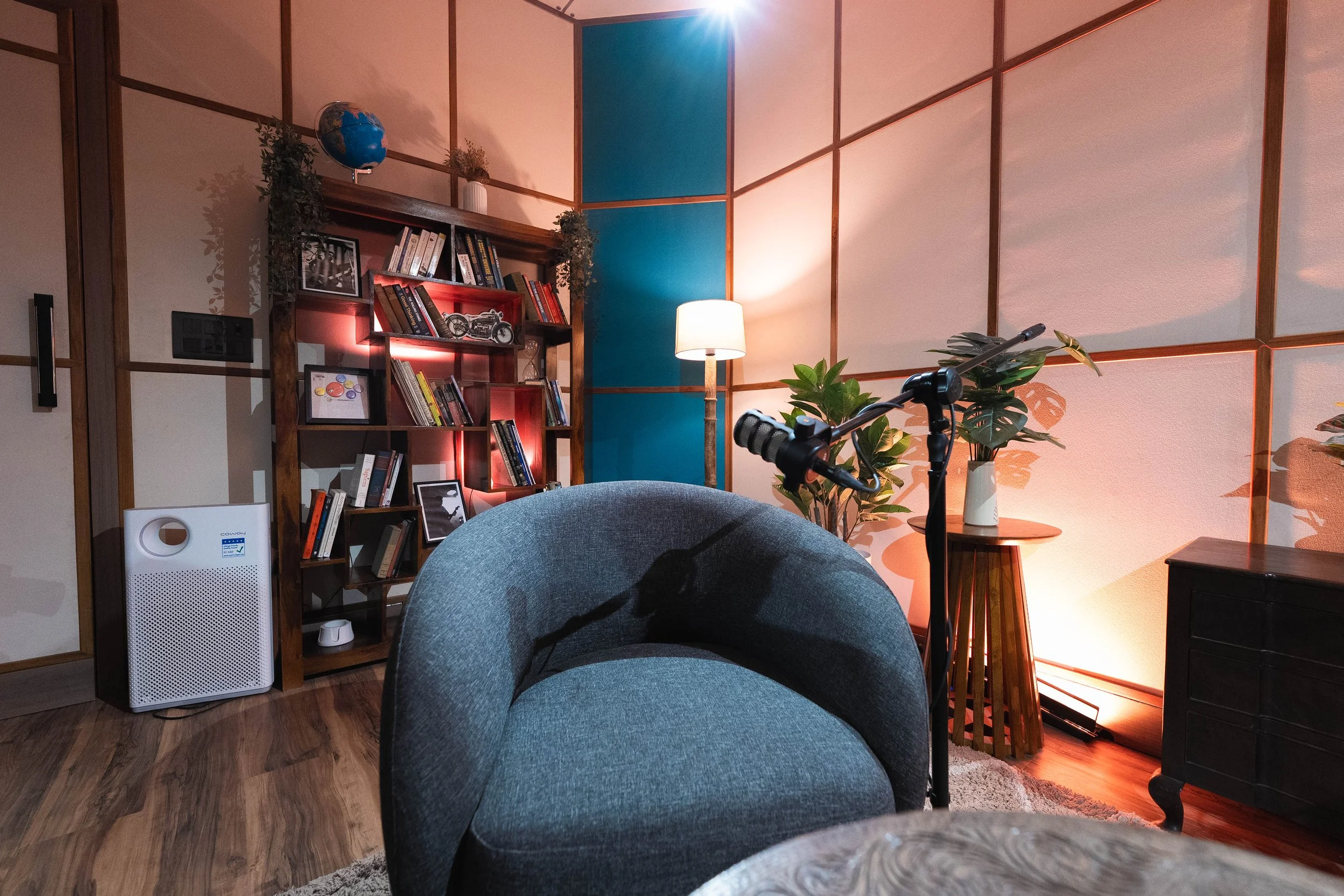 A cozy living room corner featuring a round gray armchair, a wooden side table with a vase and plant, a tall floor lamp, a blue accent wall with wood paneling, and a bookshelf with books and decor, lit by warm and ambient lighting.