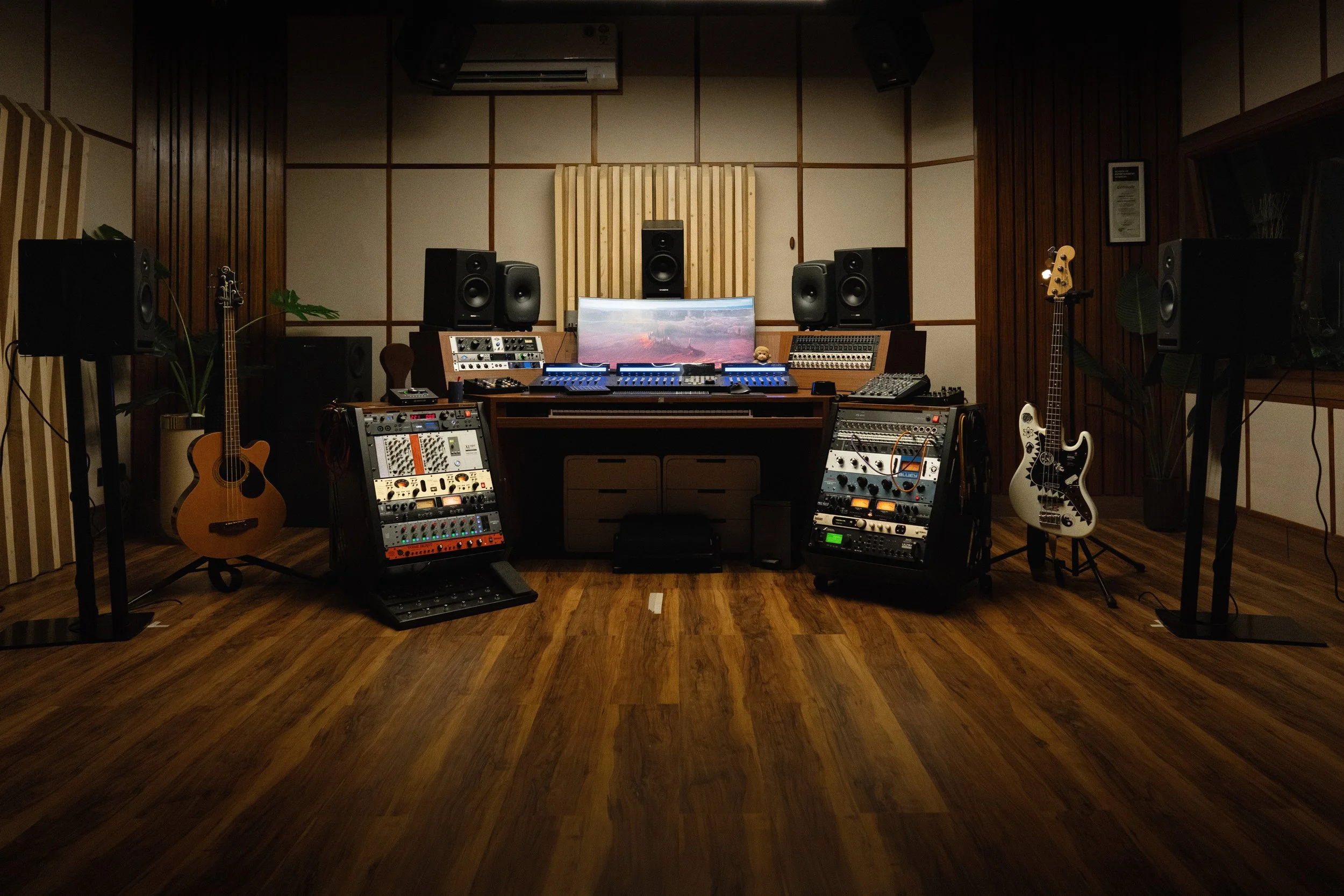 A music recording studio with various speakers, musical instruments, audio equipment, and a computer monitor on a wooden desk, set in a room with wooden walls and flooring.