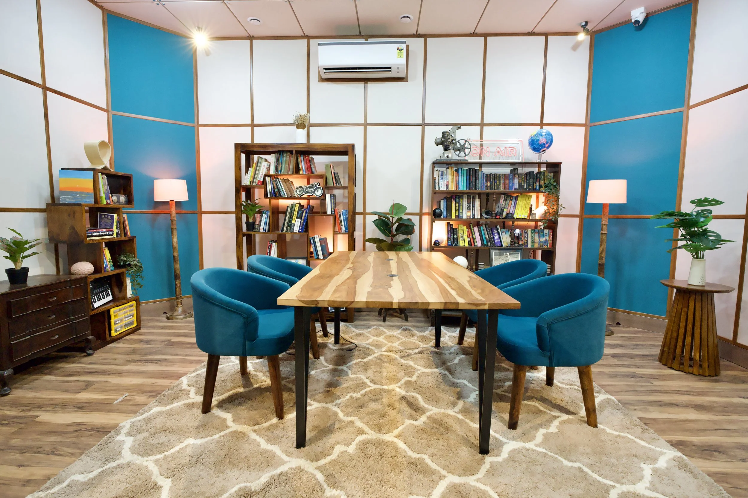 A modern conference room with a wooden table and six blue upholstered chairs. The walls feature white and blue panels with wooden framing. There are three bookshelves filled with books, a neon 'On Air' sign, a globe, and decorative plants. The room h