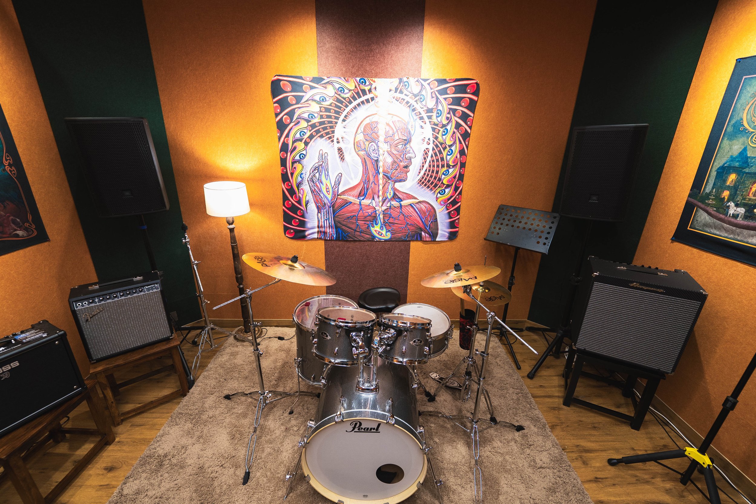 A music room with a Pearl drum set, two large PA speakers, a guitar amplifier, a music stand, two wall-mounted artworks, a floor lamp, and acoustic panels on the walls.