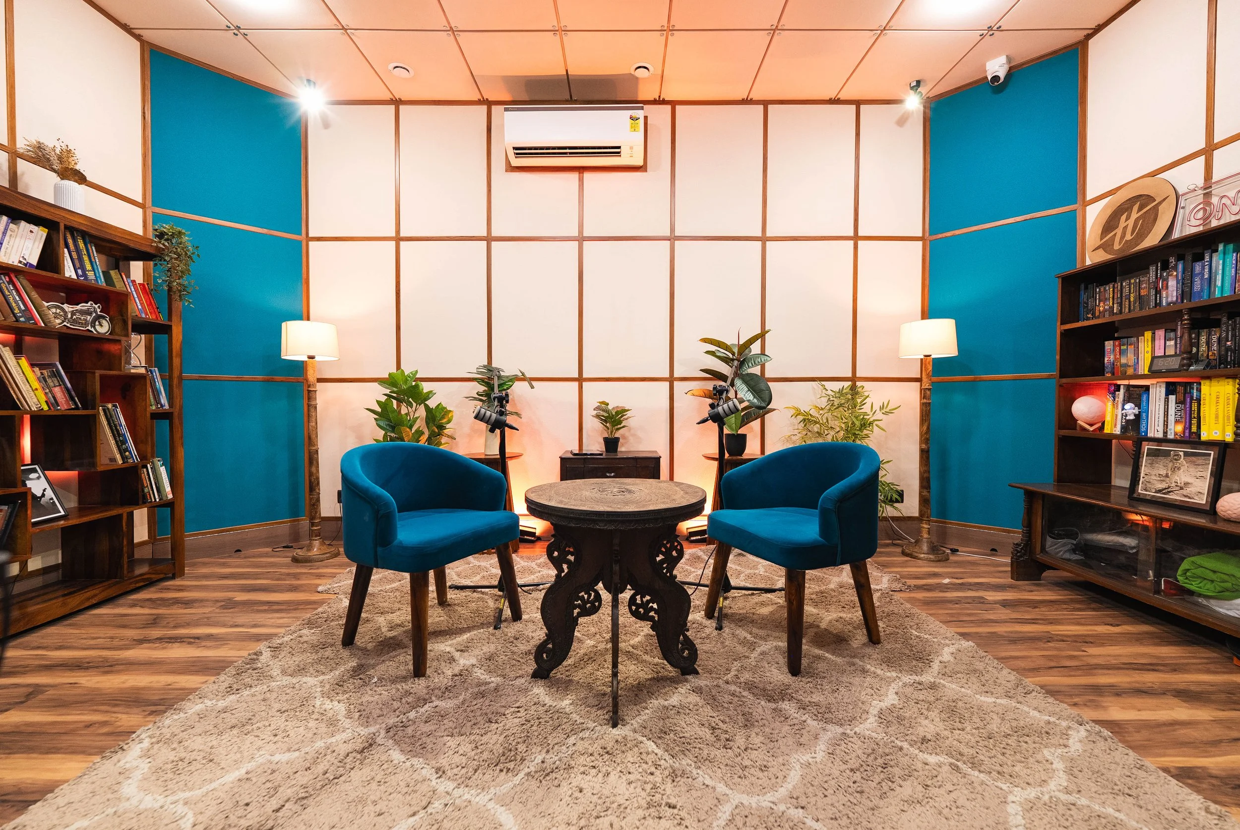 a podcast room with two chairs and a small, round table in the foreground and two bookshelves and plants in the background. 