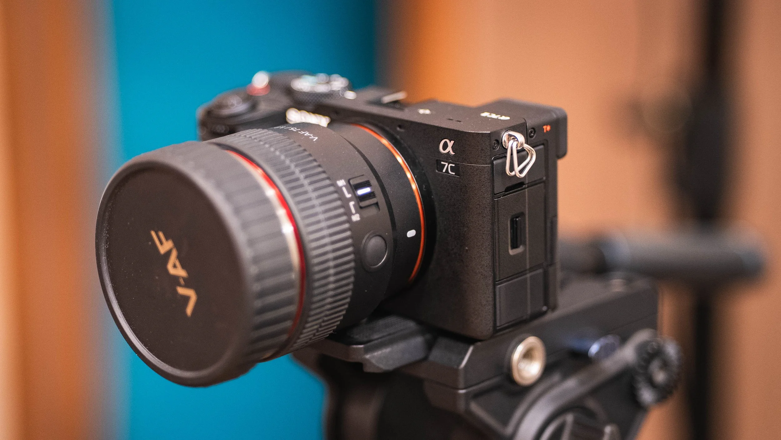 Close-up of a black mirrorless camera with a large lens, mounted on a tripod or stabilizer, blurred background with blue and orange tones.