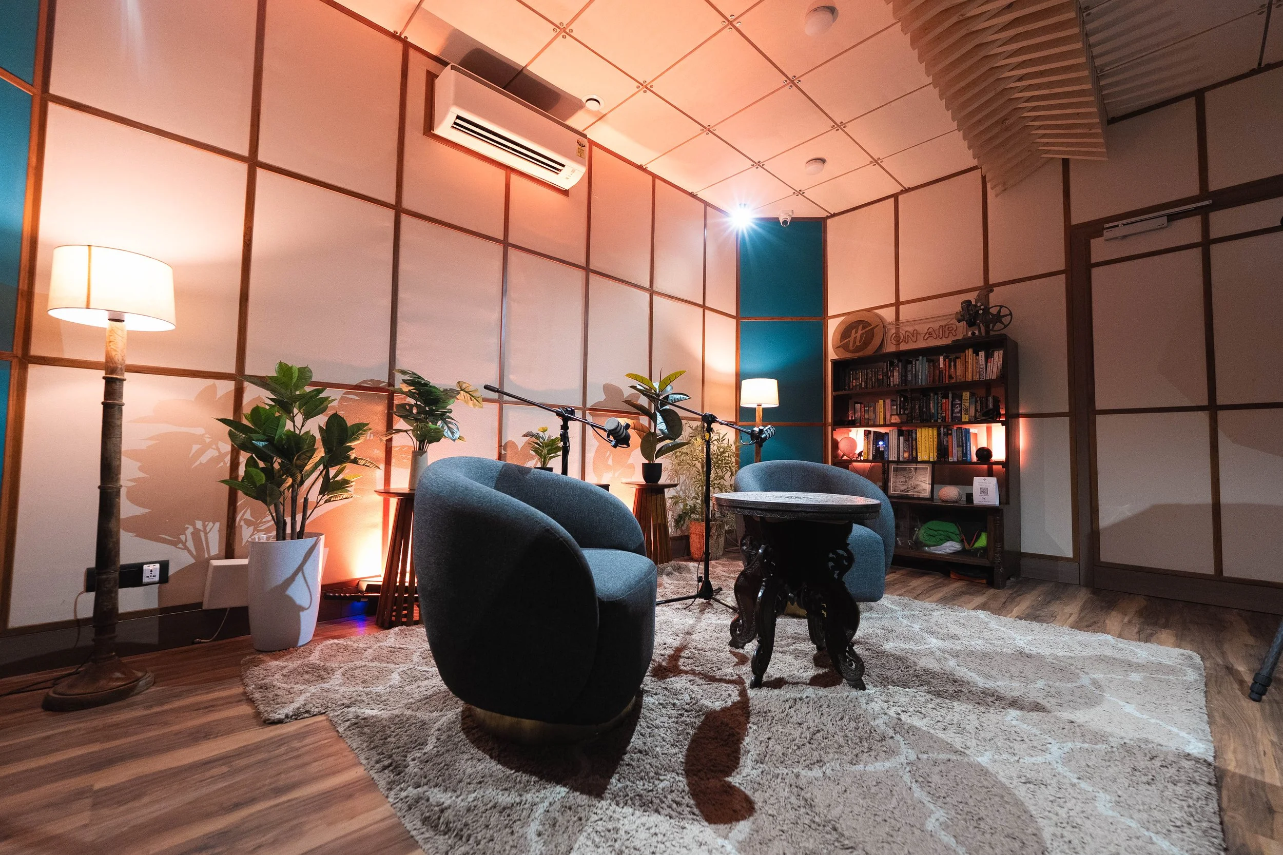 A cozy podcast recording studio with chairs, plants, a bookshelf, and warm lighting, including a floor lamp and table lamps.