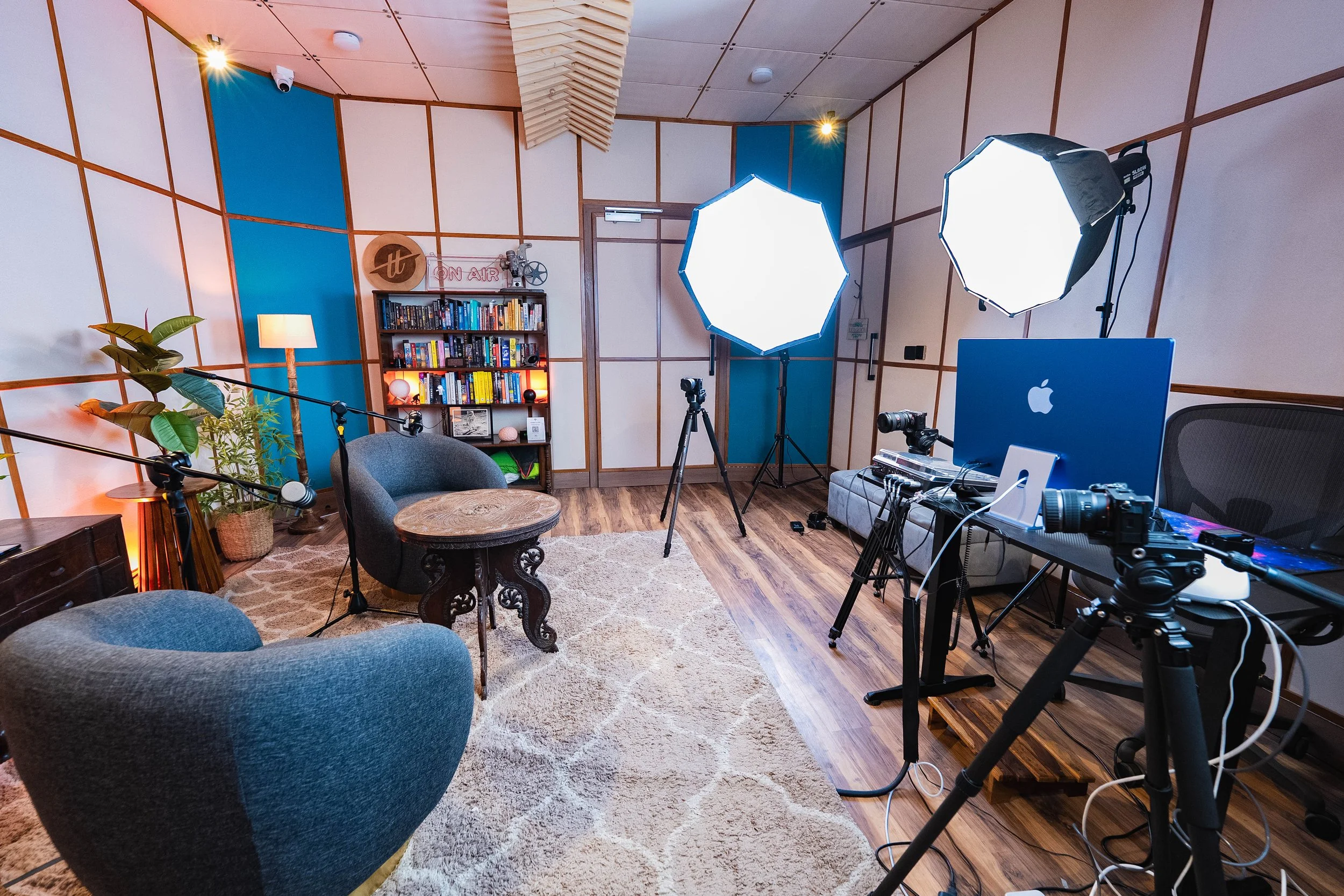 A room set up for video recording or streaming with professional lighting equipment and cameras. There are two large softbox lights directed toward the center, a camera on a tripod, a desk with a computer, and a bookshelf with books and decorative it