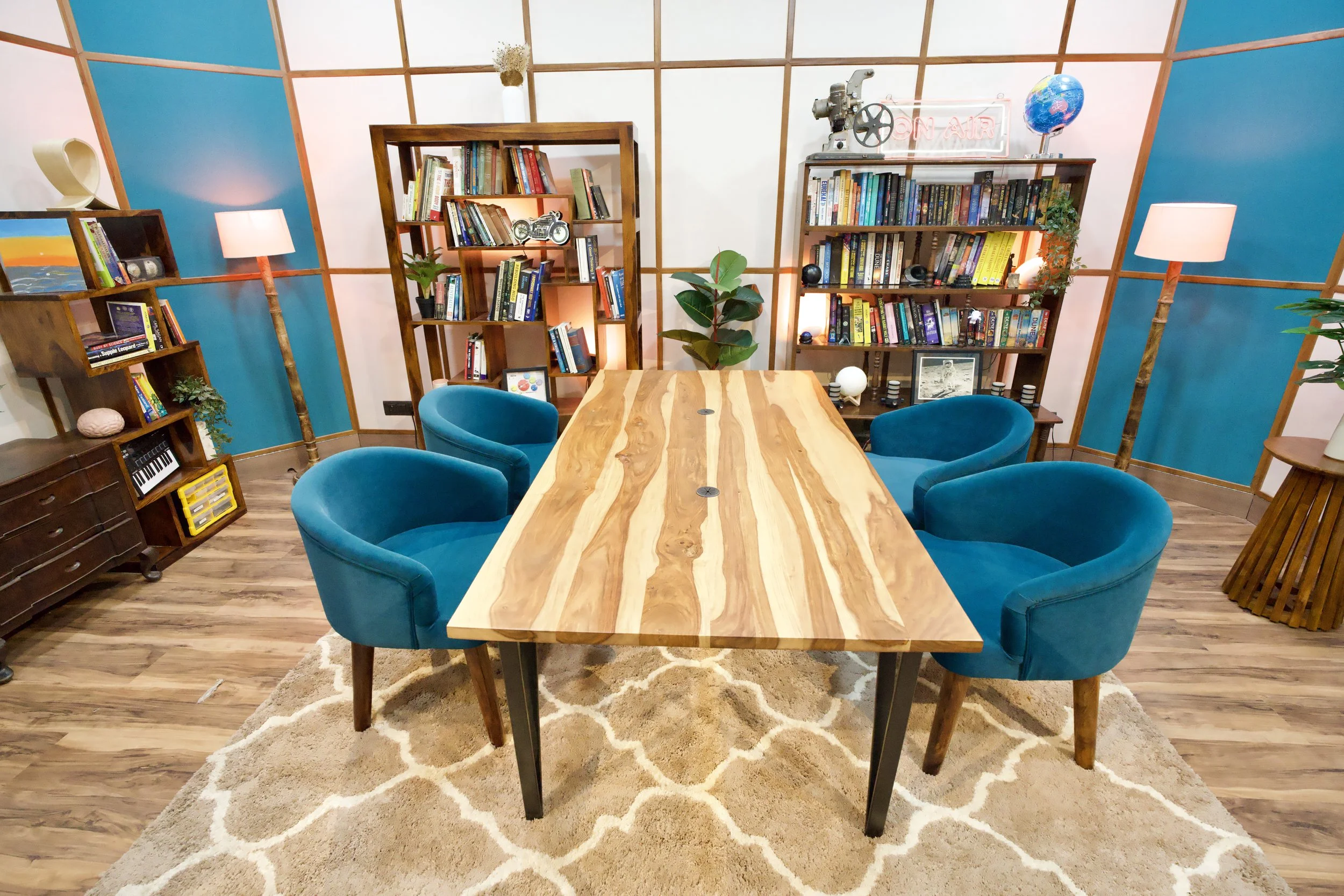 Meeting room with a wooden table, six teal velvet chairs, bookshelves filled with books, plants, lamps, and decorative items.