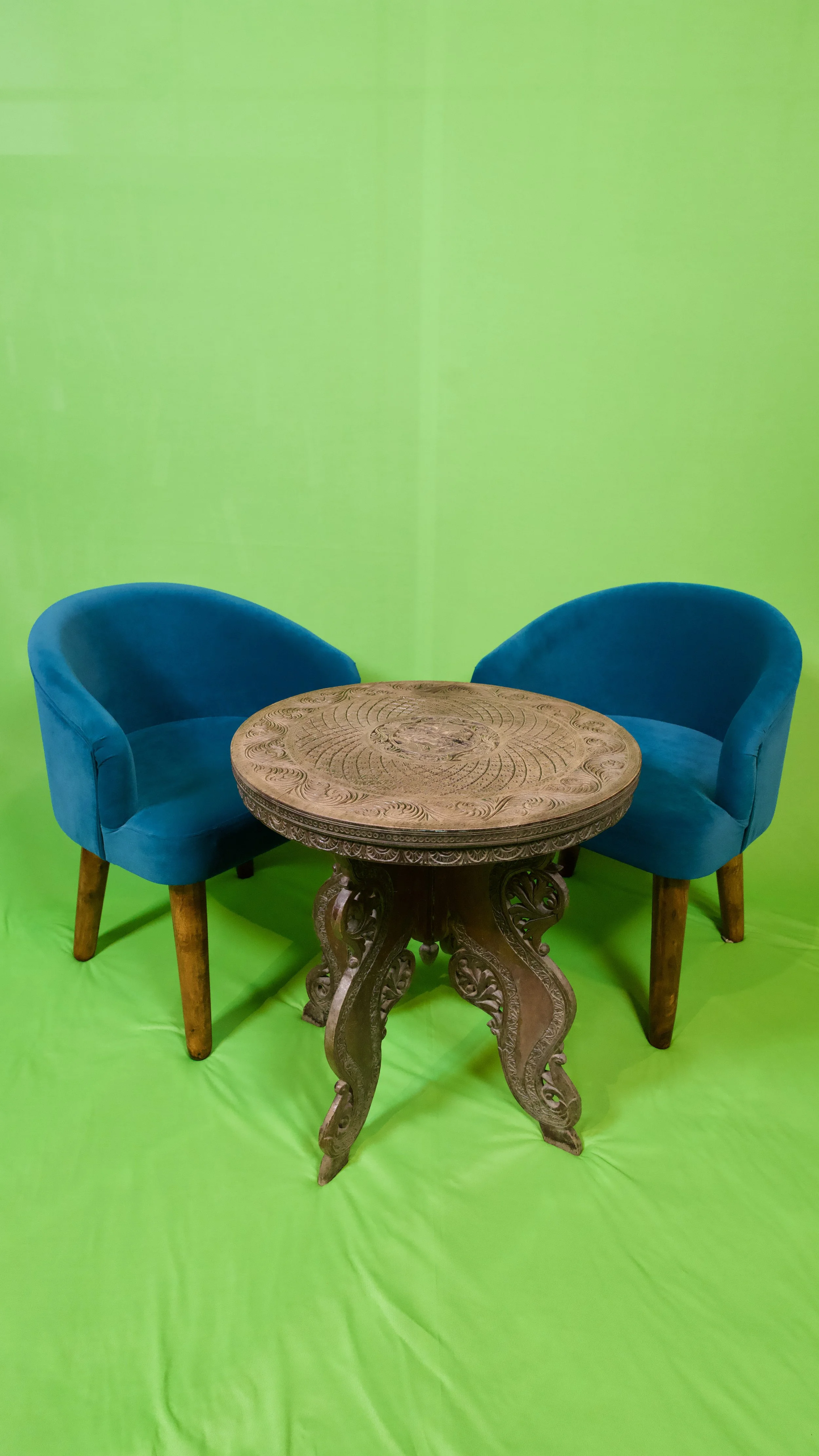 Two blue upholstered armchairs with wooden legs surrounding a carved round wooden table, set against a bright green background.