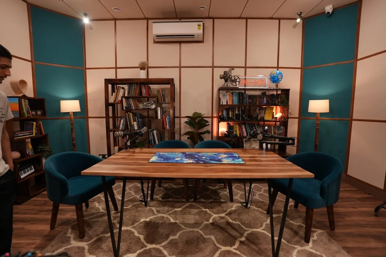 a podcast room with four chairs and a small, round table in the foreground and two bookshelves and plants in the background. 