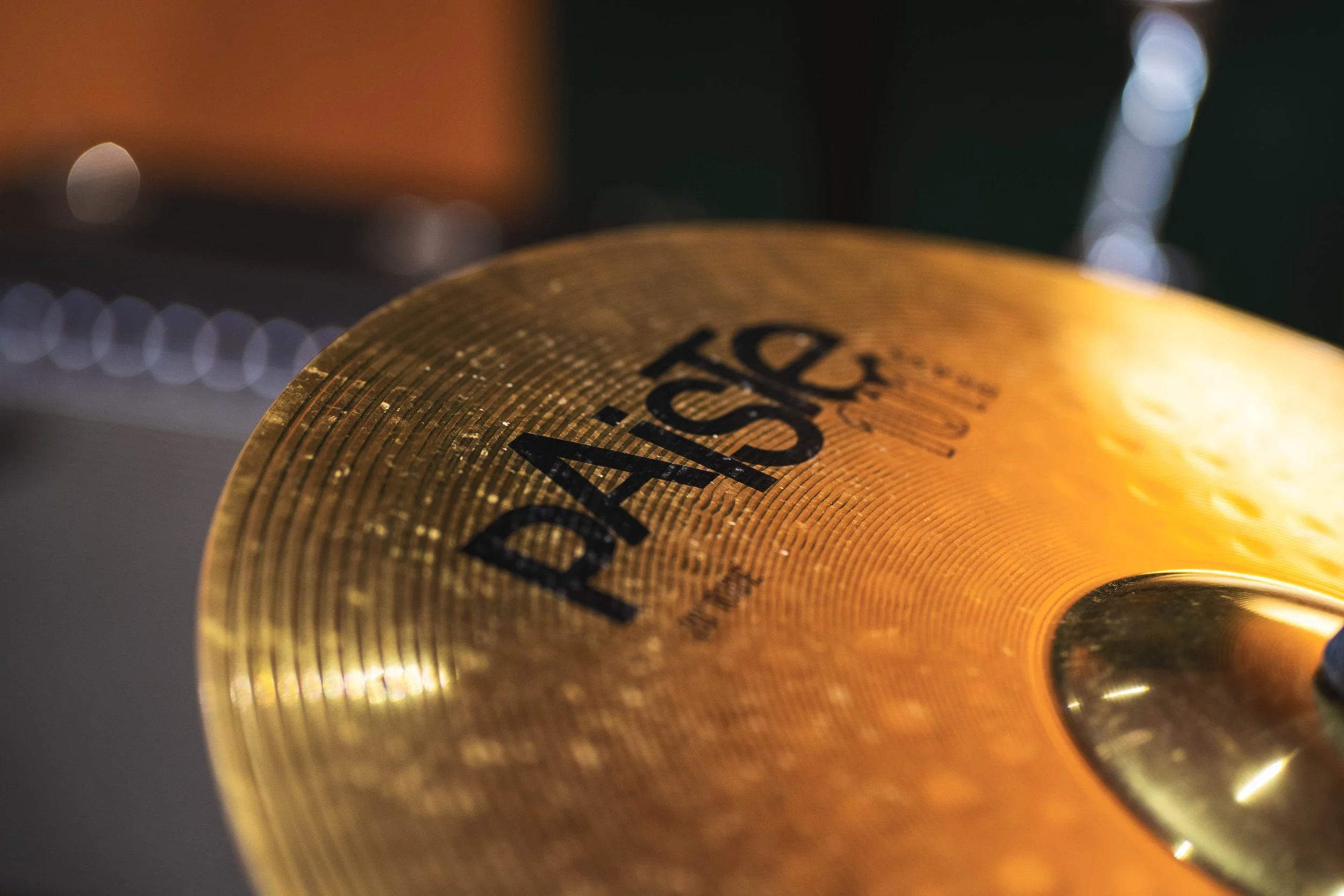 Close-up shot of a golden-colored cymbal with black text, showing the brand name 'Paiste' and part of the musical instrument's surface with a textured pattern.