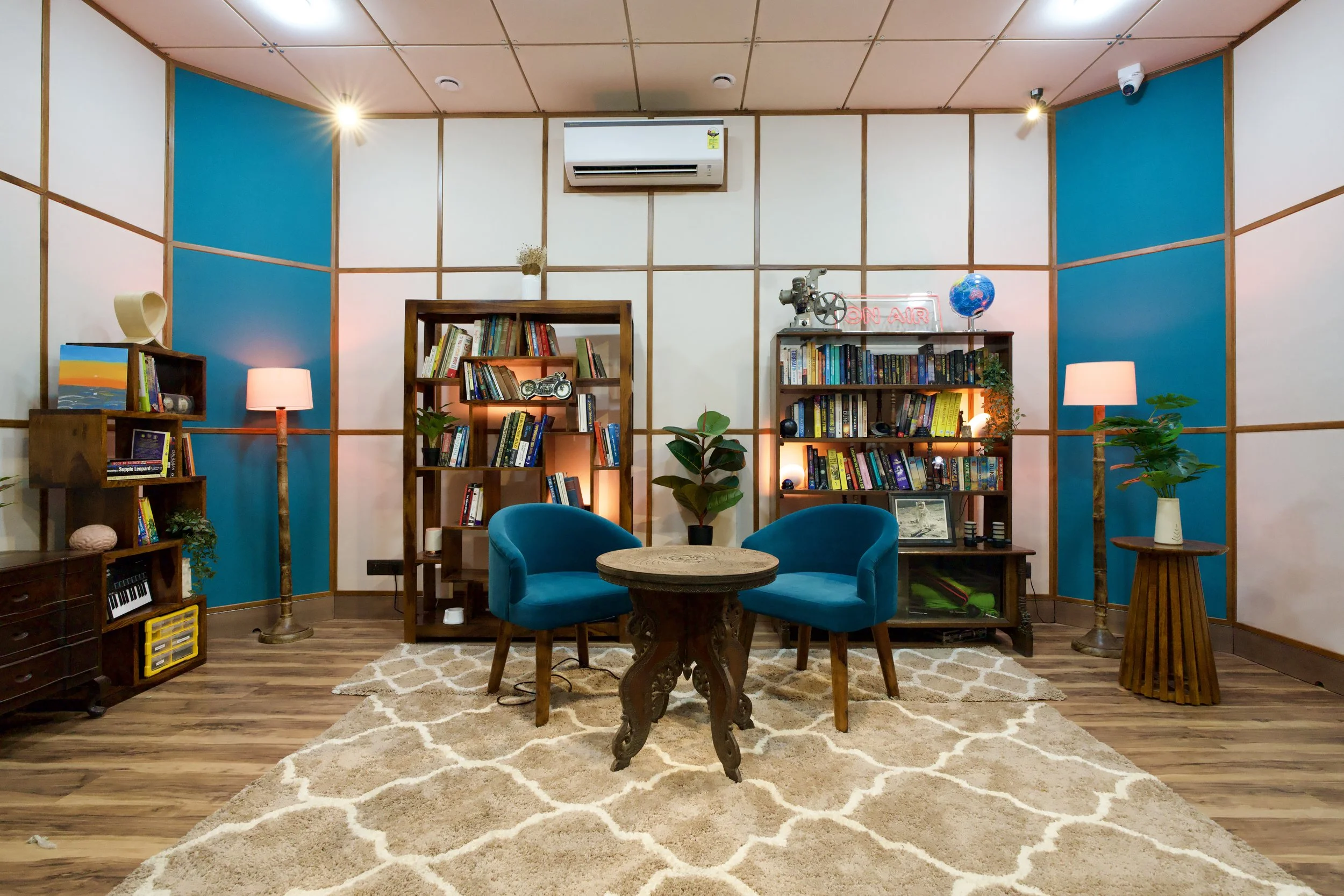 A cozy room with wooden floors, shelving filled with books, two blue armchairs around a small round wooden table, and two floor lamps with pink shades. The walls feature large white and blue panels, with a wall-mounted air conditioner and a neon sign