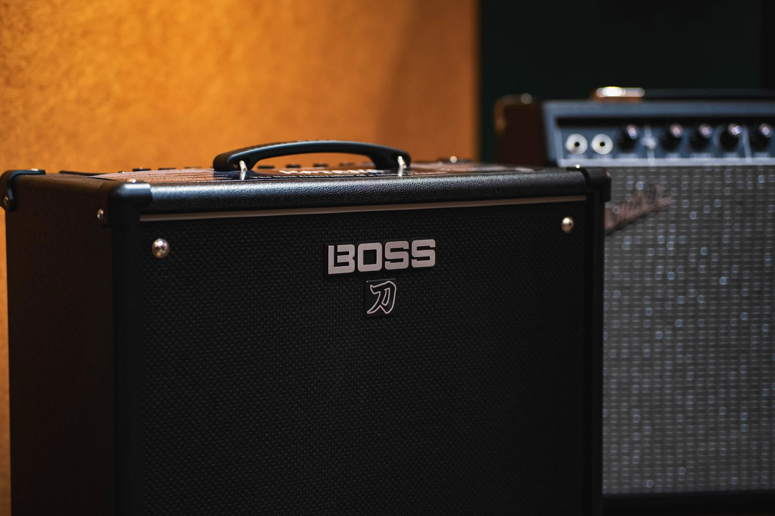 Close-up of a black BOSS guitar amplifier with a textured surface, silver logo, and a Fender amplifier in the background.