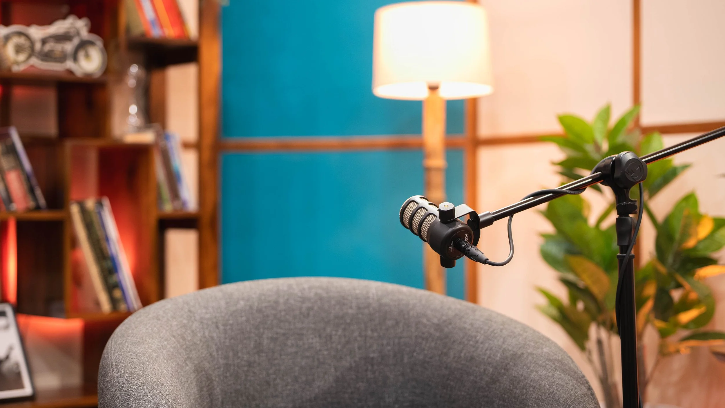 A microphone on a boom arm in an indoor setting with a gray chair in the foreground, a bookshelf with books and decorative items, a floor lamp, and green plants in the background.