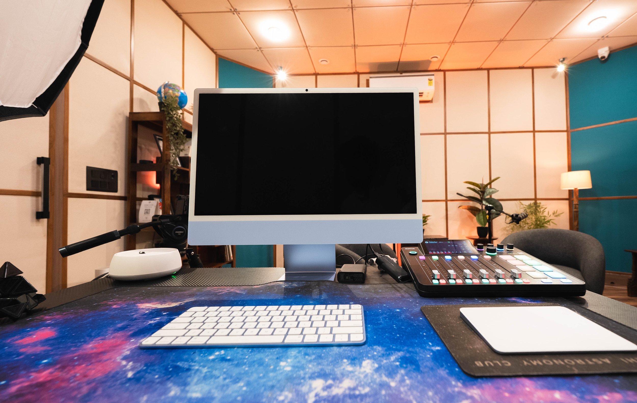 Music and video production setup with a computer monitor, keyboard, mixing controller, microphone, and studio lighting in a room with wooden paneling and plants.