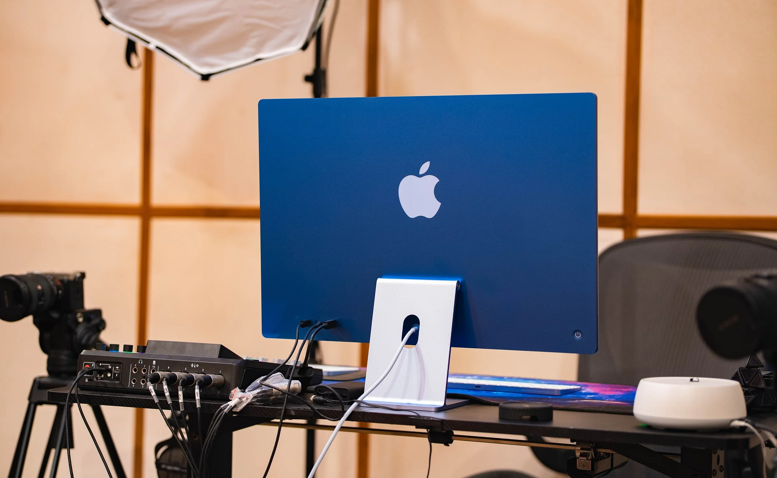 A workstation with a blue Apple monitor, connected to various audio and video equipment, on a black desk in a studio setting.