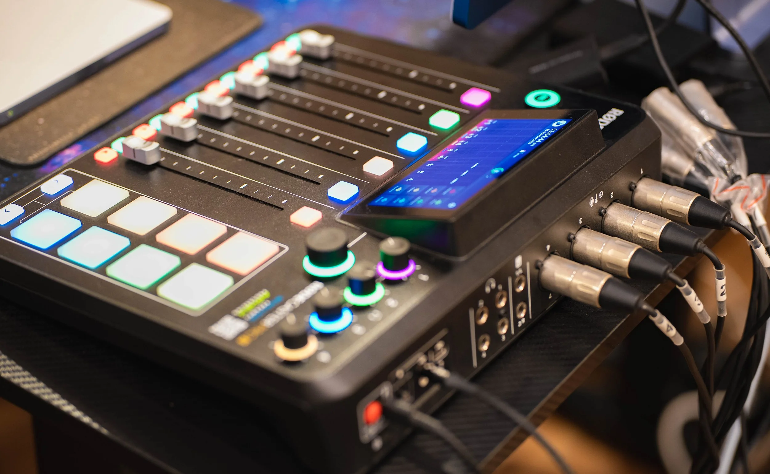 A music mixing console with colorful illuminated buttons and faders, connected to multiple cables.