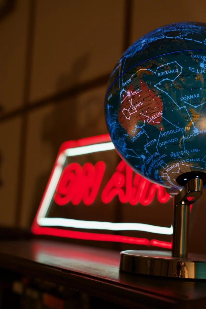 A globe showing a map of Australia with labeled regions, placed on a desk with a blurred neon sign reading 'OPEN' in the background.