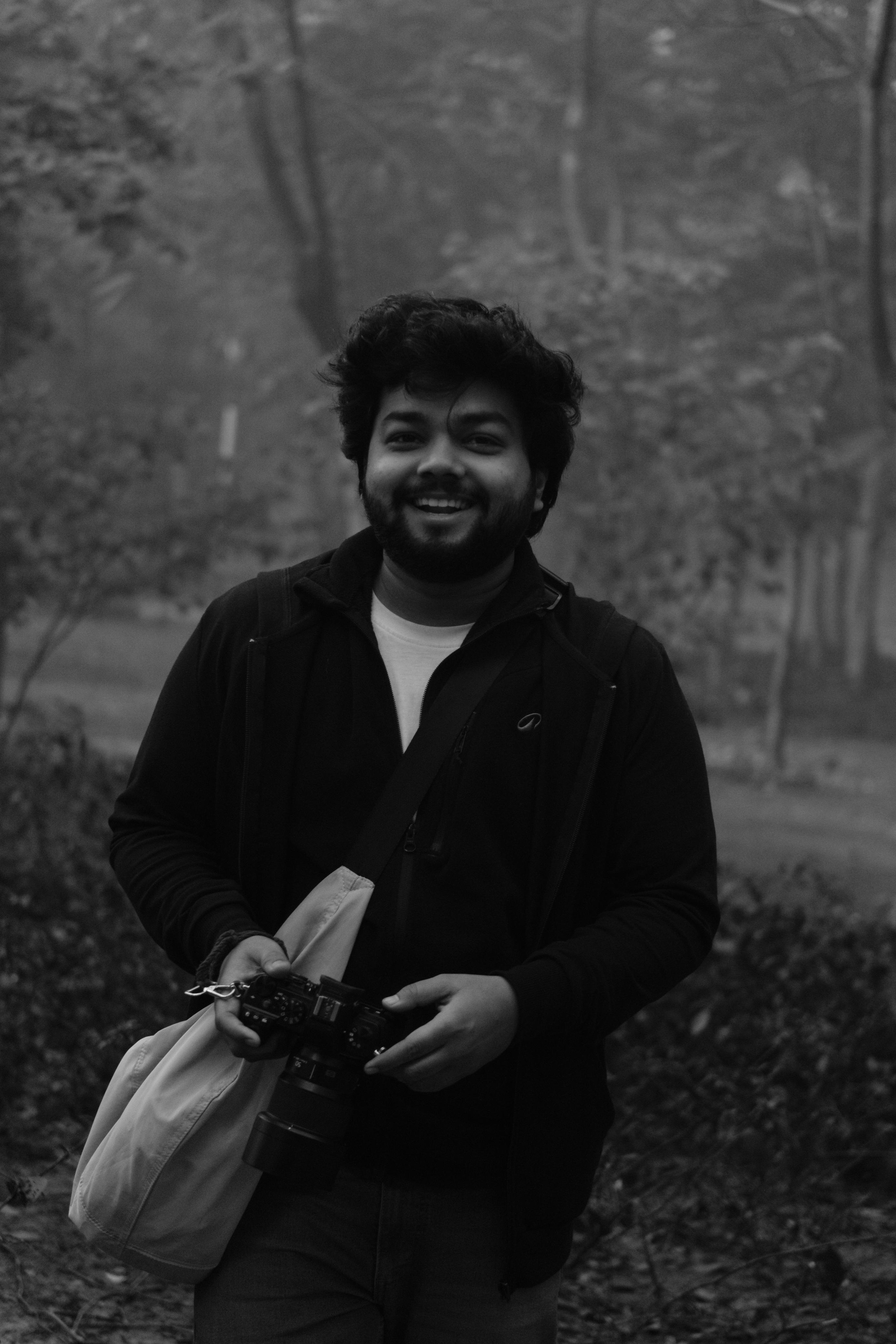 A man with curly dark hair and a beard is smiling outdoors in a forested area, holding a camera and a beige bag, wearing a dark jacket.