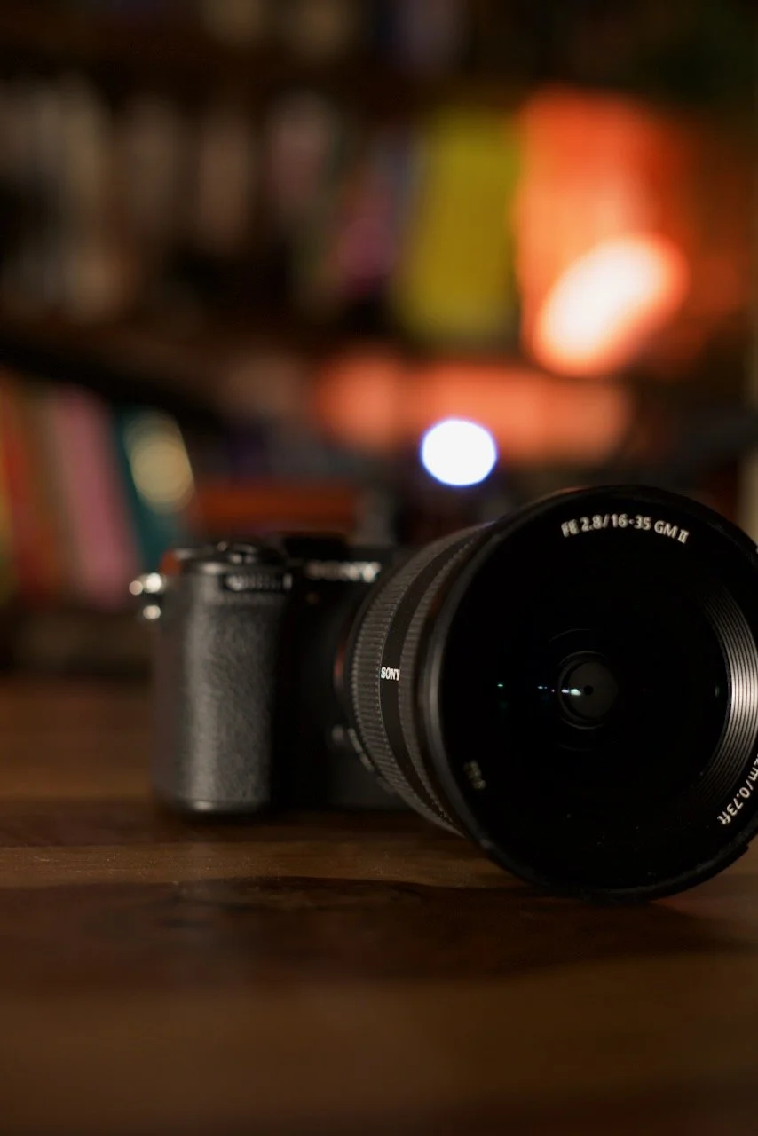 A camera with a large zoom lens resting on a wooden surface with a blurred background of books and colorful objects.