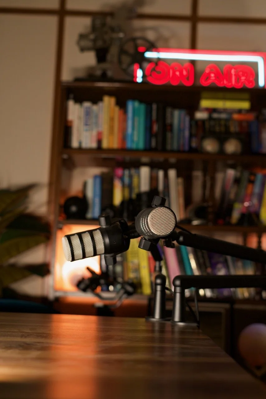 A close-up of two microphones on a wooden desk in a podcast or recording studio, with a bookshelf filled with books and a neon 