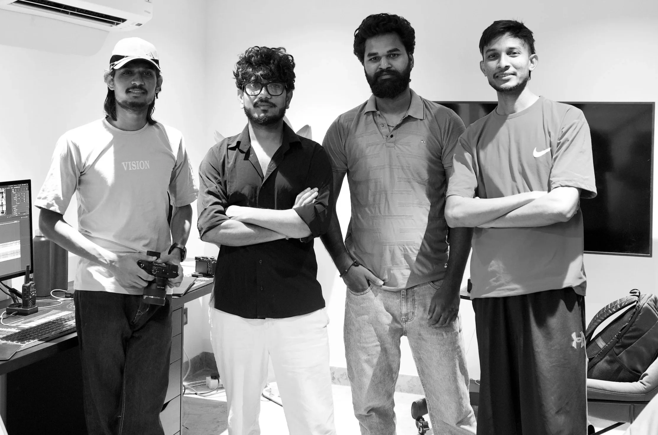 Four young men standing in a well lit room with a computer, a split air conditioner and a TV in the background. 