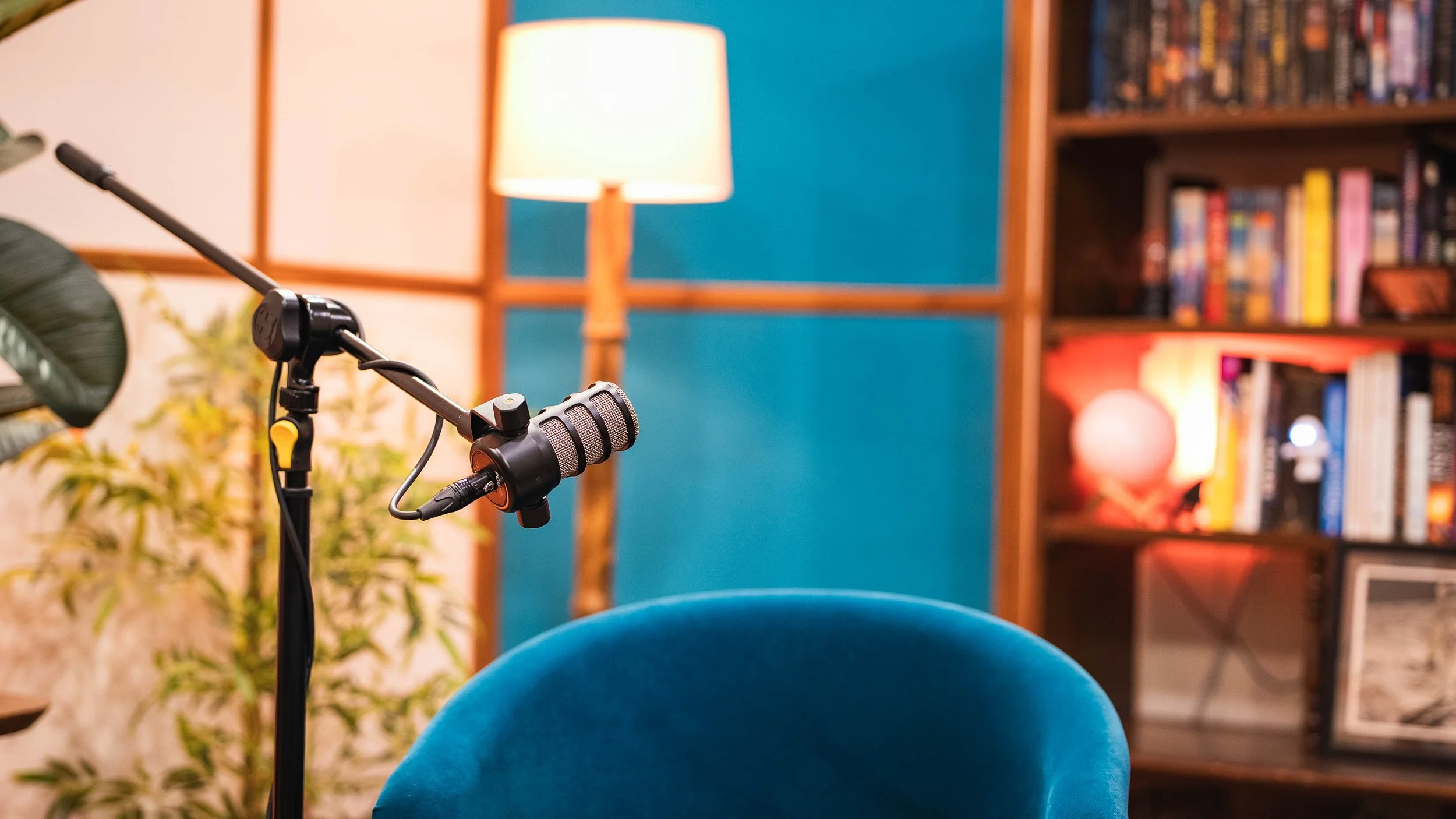 A microphone on a stand in front of a blue chair, with a bookshelf, a lamp, and a plant in the background.