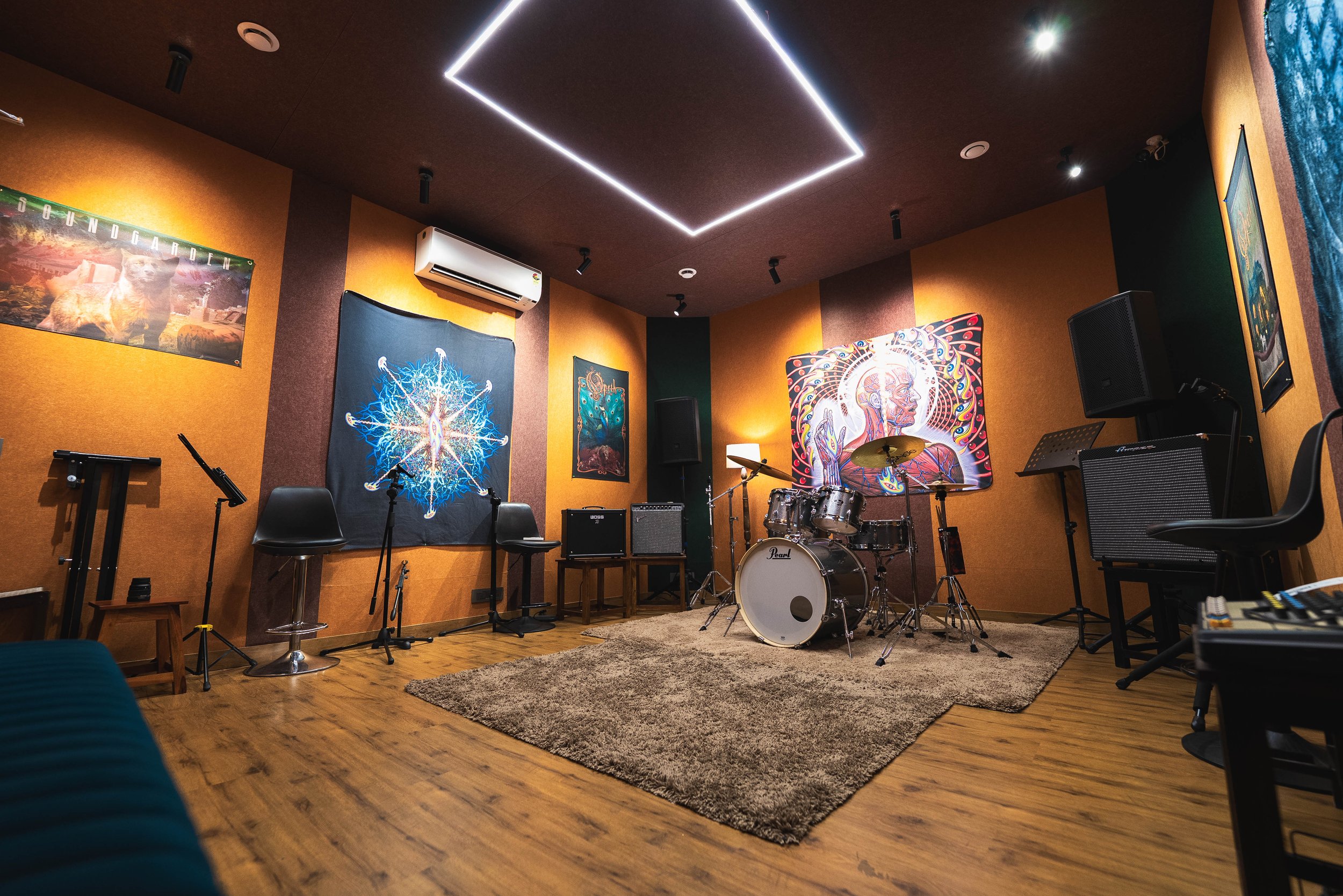 A music rehearsal room with orange and brown walls, decorated with psychedelic and abstract artwork. The room features a drum set, an amplifier, a keyboard, black chairs, and a rug on a wooden floor, with a ceiling fitted with modern LED lighting.