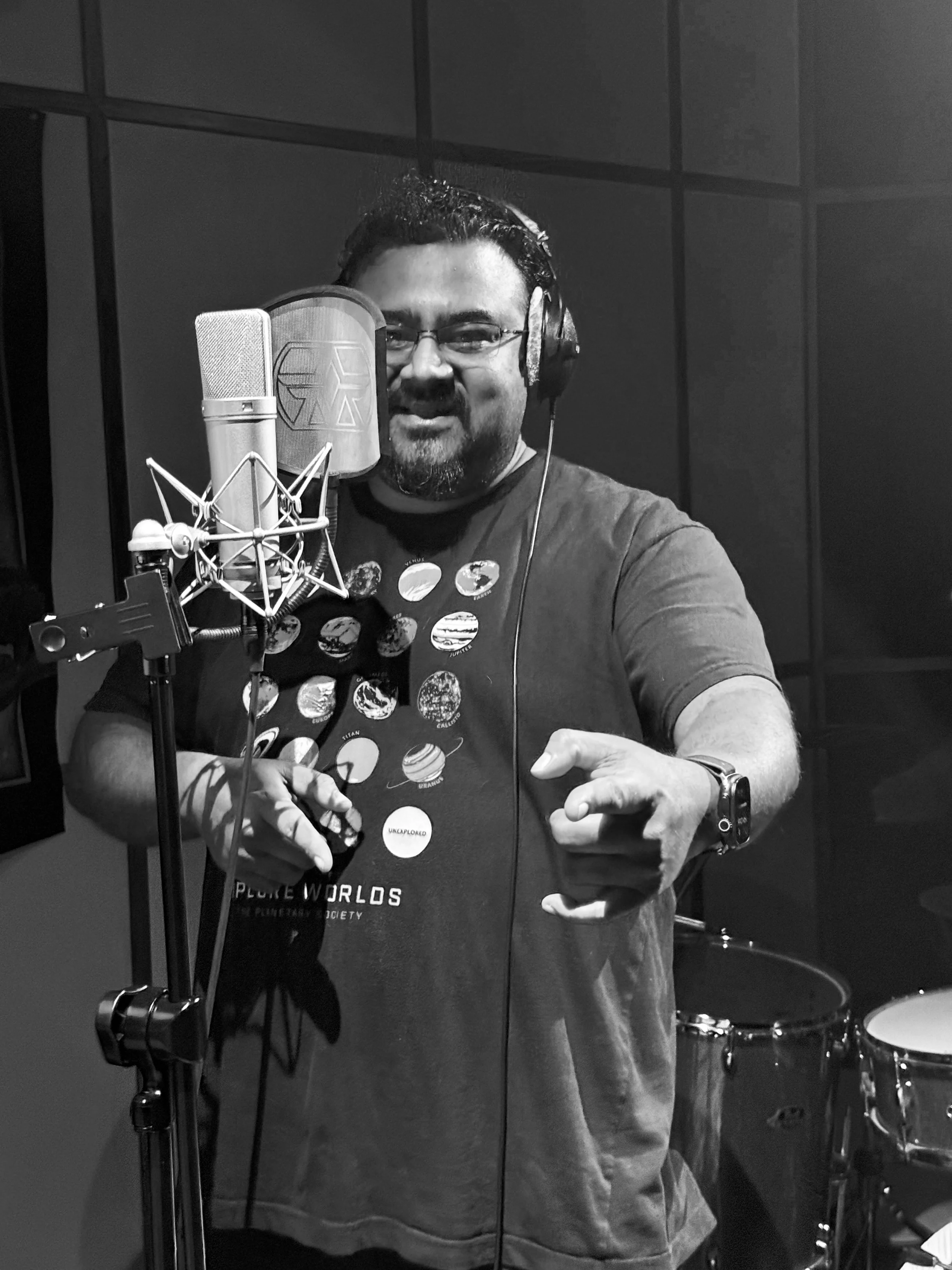 A man wearing glasses, headphones, and a t-shirt with planets printed on it, standing in a recording studio in front of a microphone and pop filter, smiling at the camera.