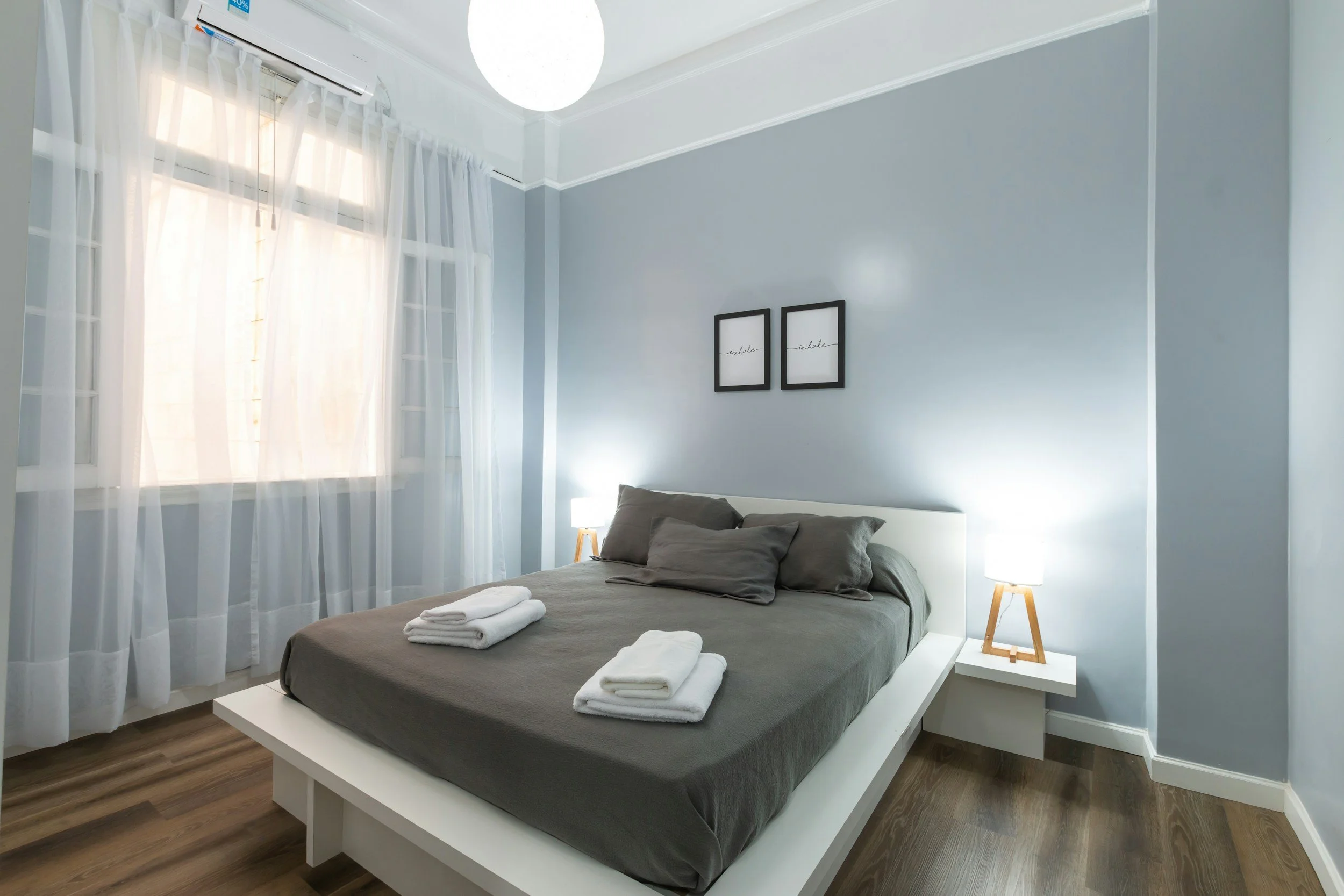 Minimalist bedroom with a gray bed, matching pillows and folded towels, beside white floating nightstands with table lamps, light blue walls, sheer white curtains, and a window with an air conditioning unit.