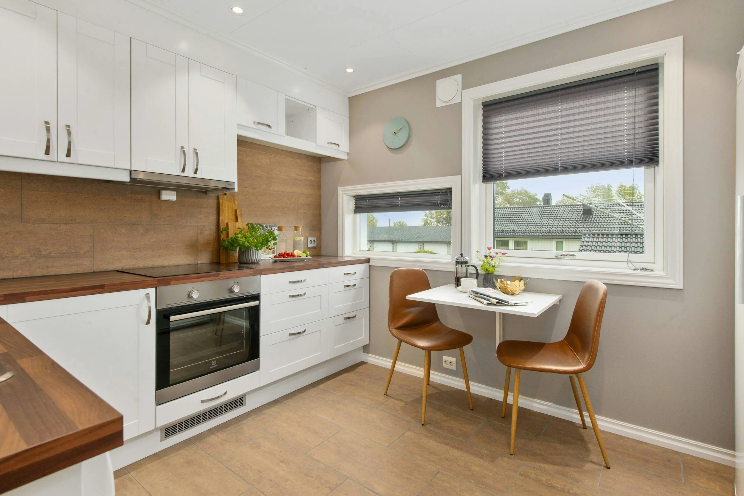 Kitchen with white cabinets, wooden countertops, and two windows with gray blinds. There are two brown chairs and a small table with breakfast items near the windows.