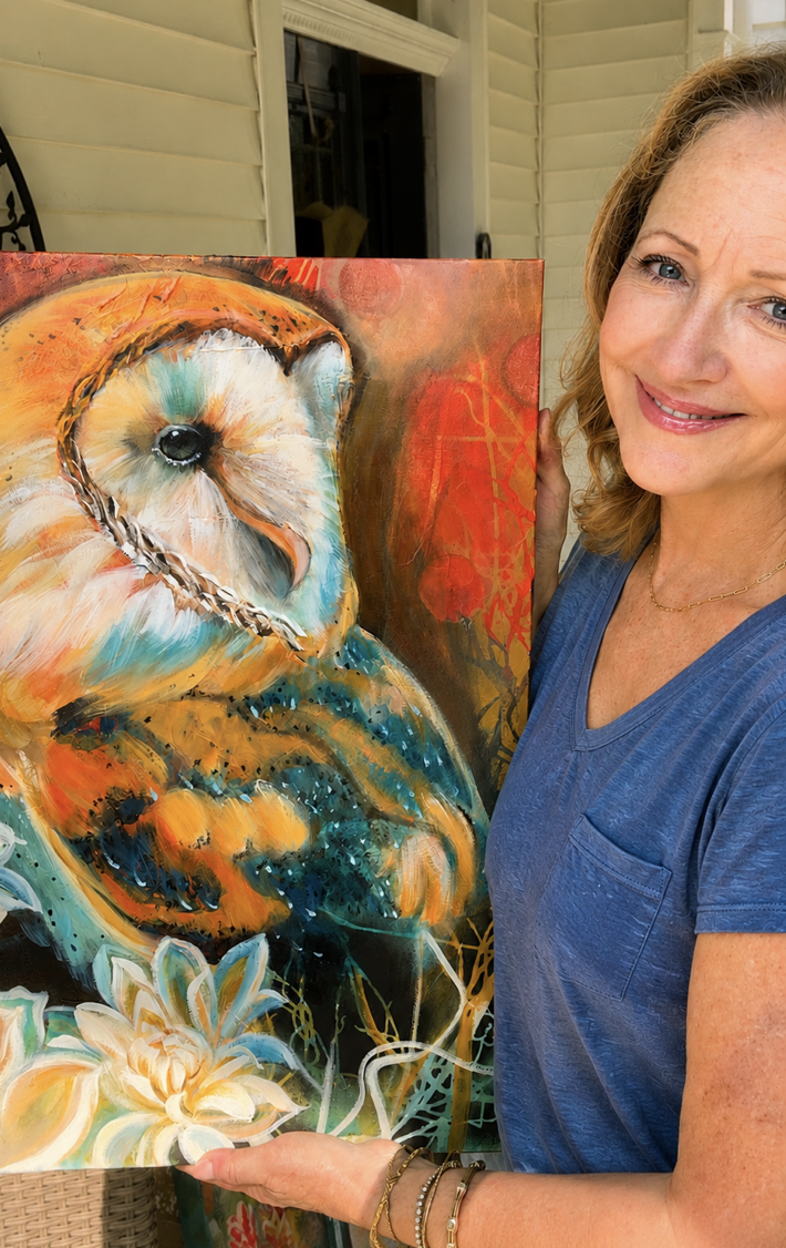 A woman with blonde hair smiling and holding a colorful painting of an owl in front of her. The painting features an abstract background with bright colors and detailed owl feathers.