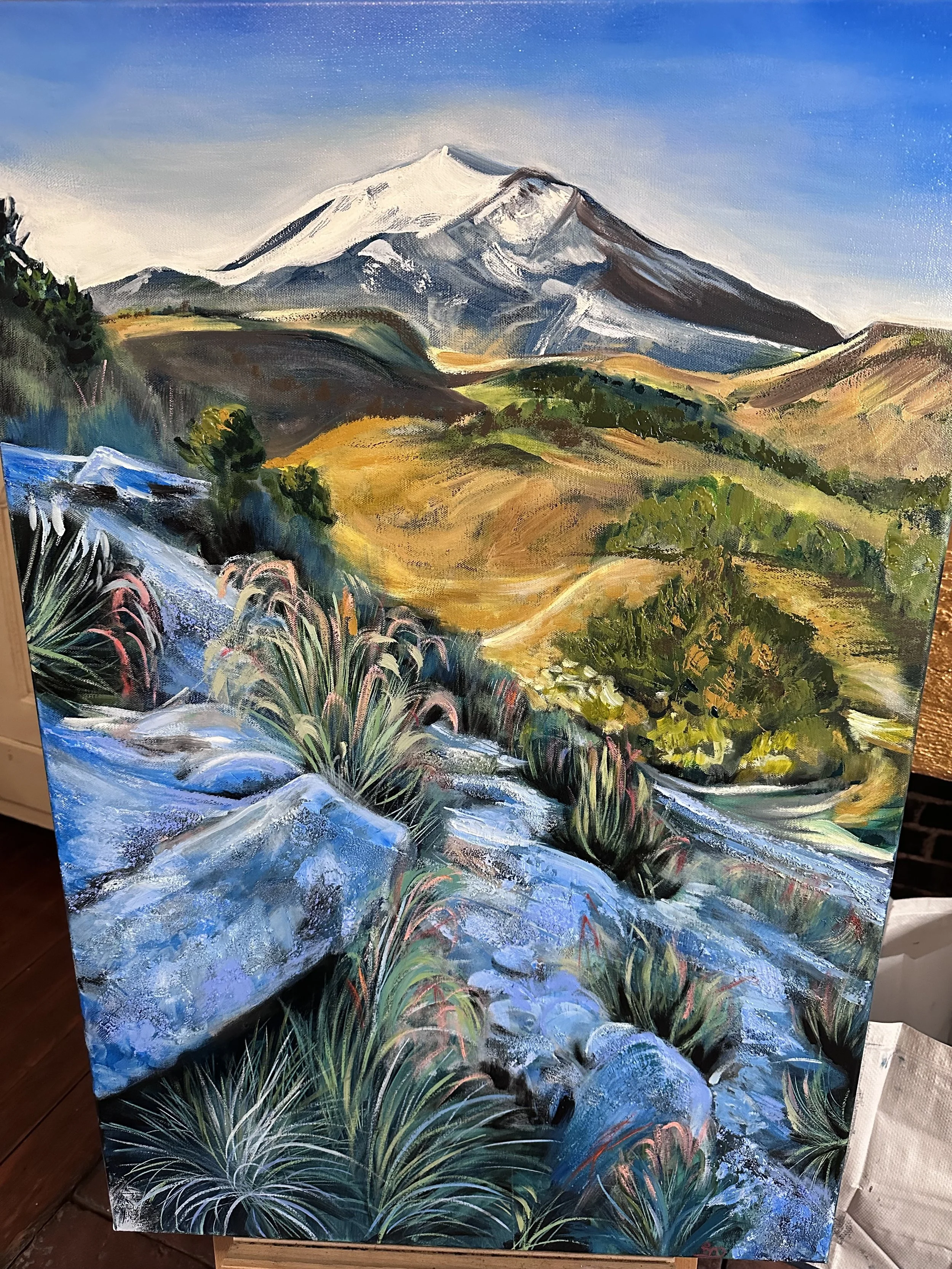 An oil painting of a snow-capped mountain in the background, with rolling green and yellow hills, and a river flowing through lush green vegetation in the foreground.