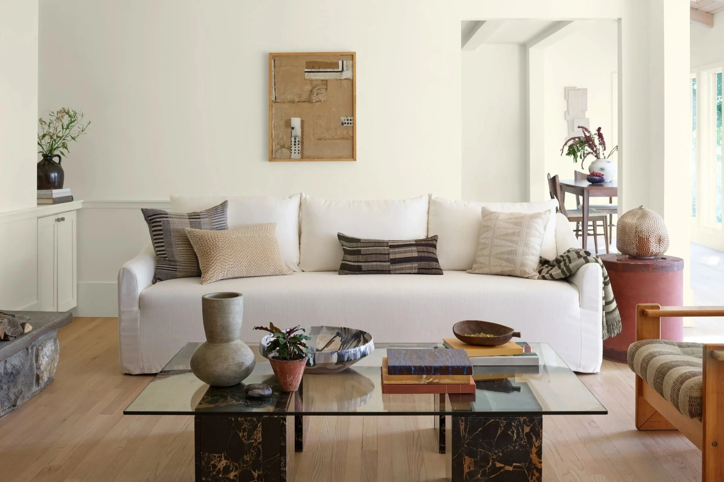 Salon moderne avec un canapé blanc, plusieurs coussins, une table basse en marbre noir, décorations dans des vases, et une fresque murale abstraite suspendue. À droite, une table de salle à manger avec un vase de fleurs, dans une pièce lumineuse avec grandes fenêtres.