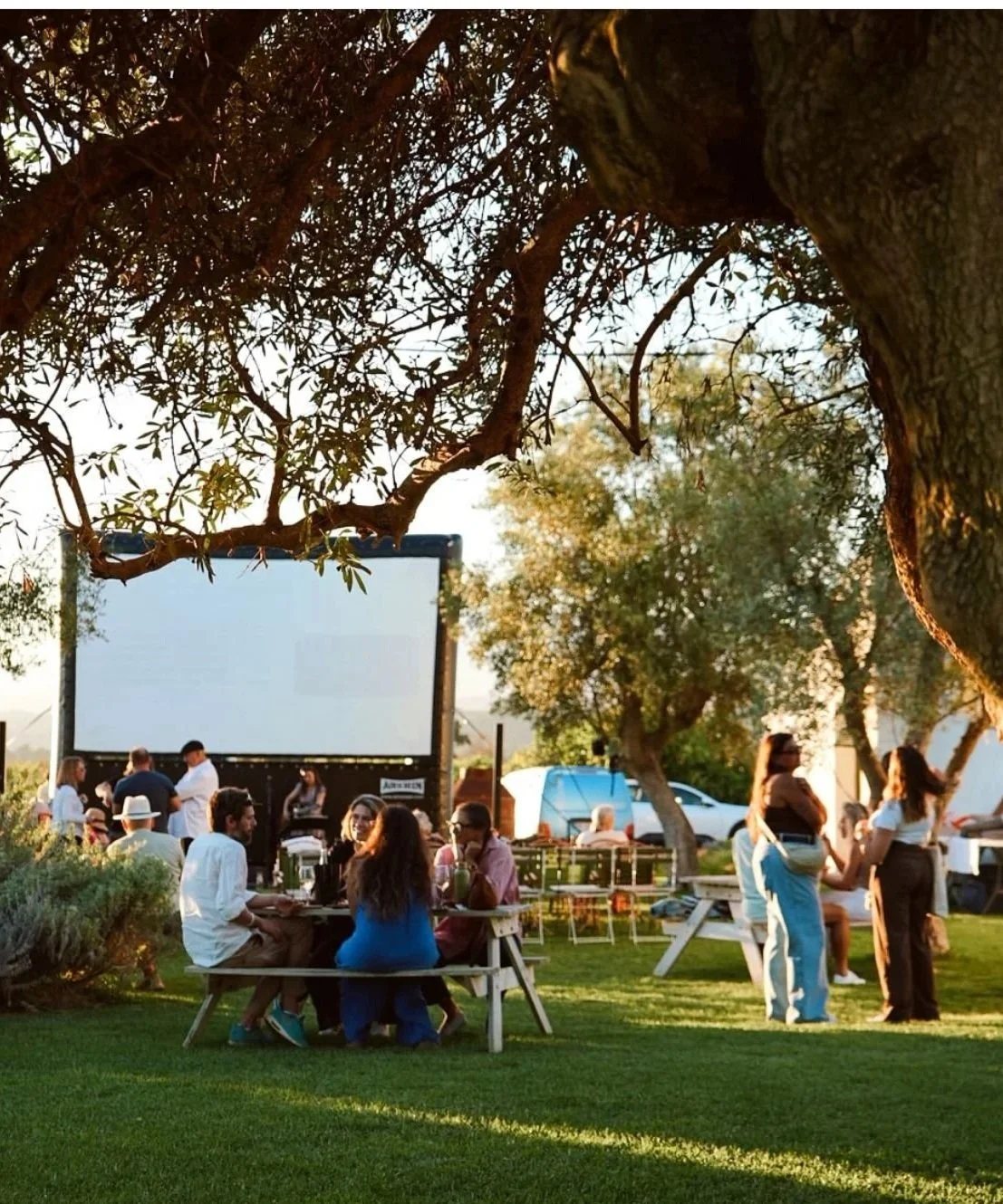 People gathering outdoors around a large outdoor movie screen, sitting on picnic tables and standing, with trees and cars in the background, during sunset.