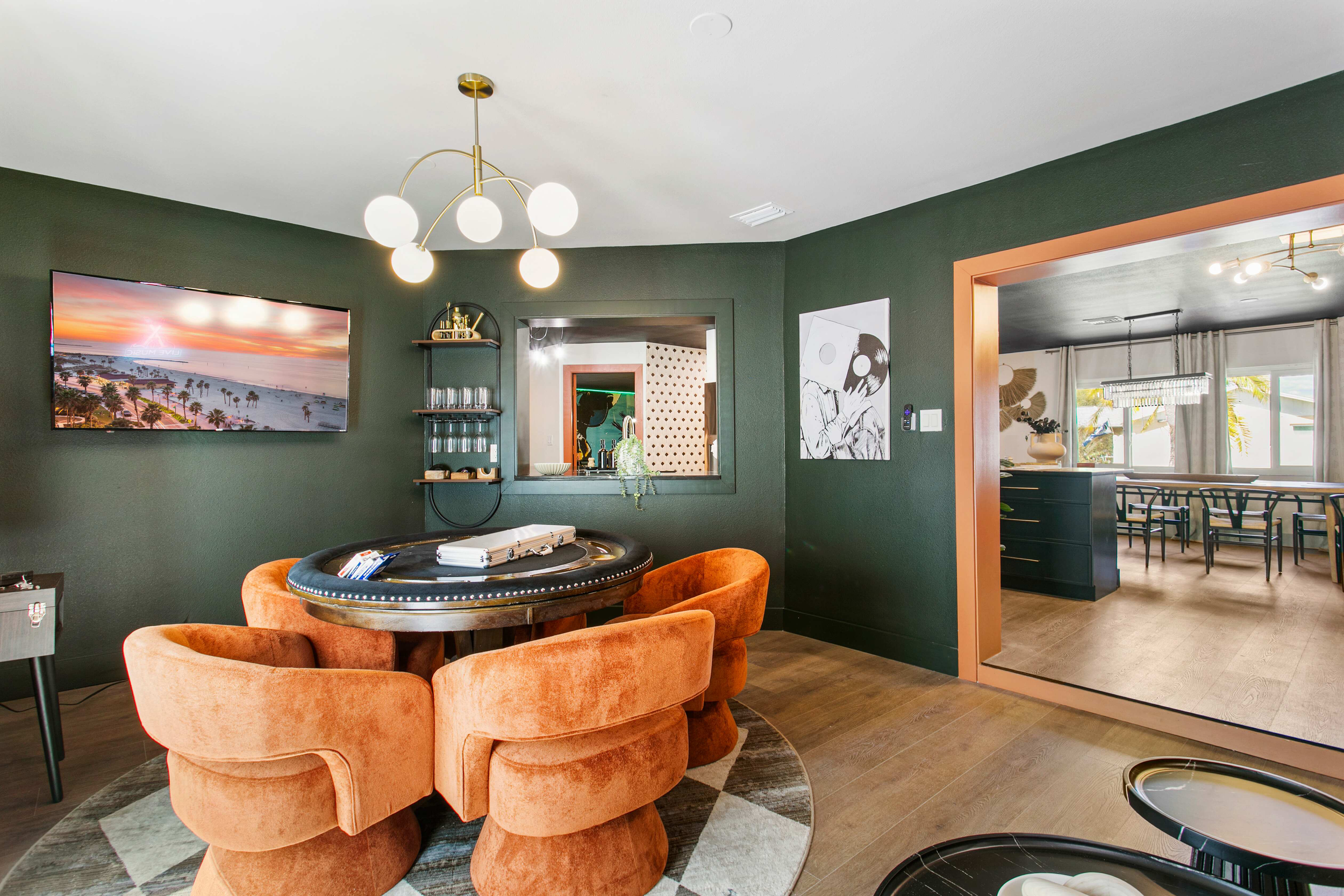 Elegant dining room with dark green walls, orange velvet chairs around a black poker-style table, and a modern chandelier with globe lights. There is a TV displaying a sunset, a bar window, and an adjacent room with a long dining table and large wind