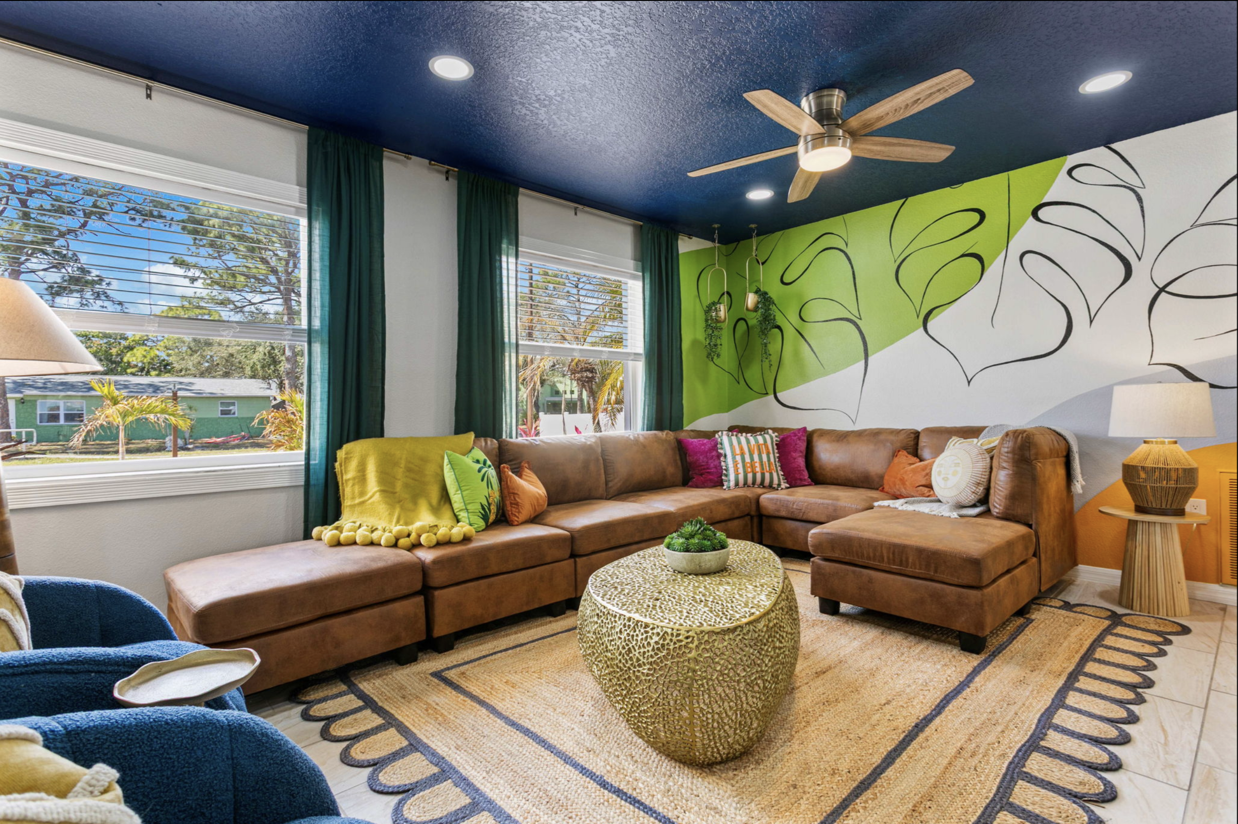 Living room with a large brown sectional sofa, colorful cushions, and a round woven coffee table on a beige rug. There are two windows with green curtains, a blue ceiling with a ceiling fan, and a wall mural with black line drawings of leaves on a gr