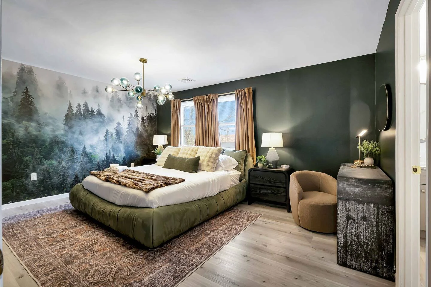 Modern bedroom with forest mural wall, king-sized bed with green velvet base, beige and green pillows, black nightstands, lamps, beige chair, and textured dresser.