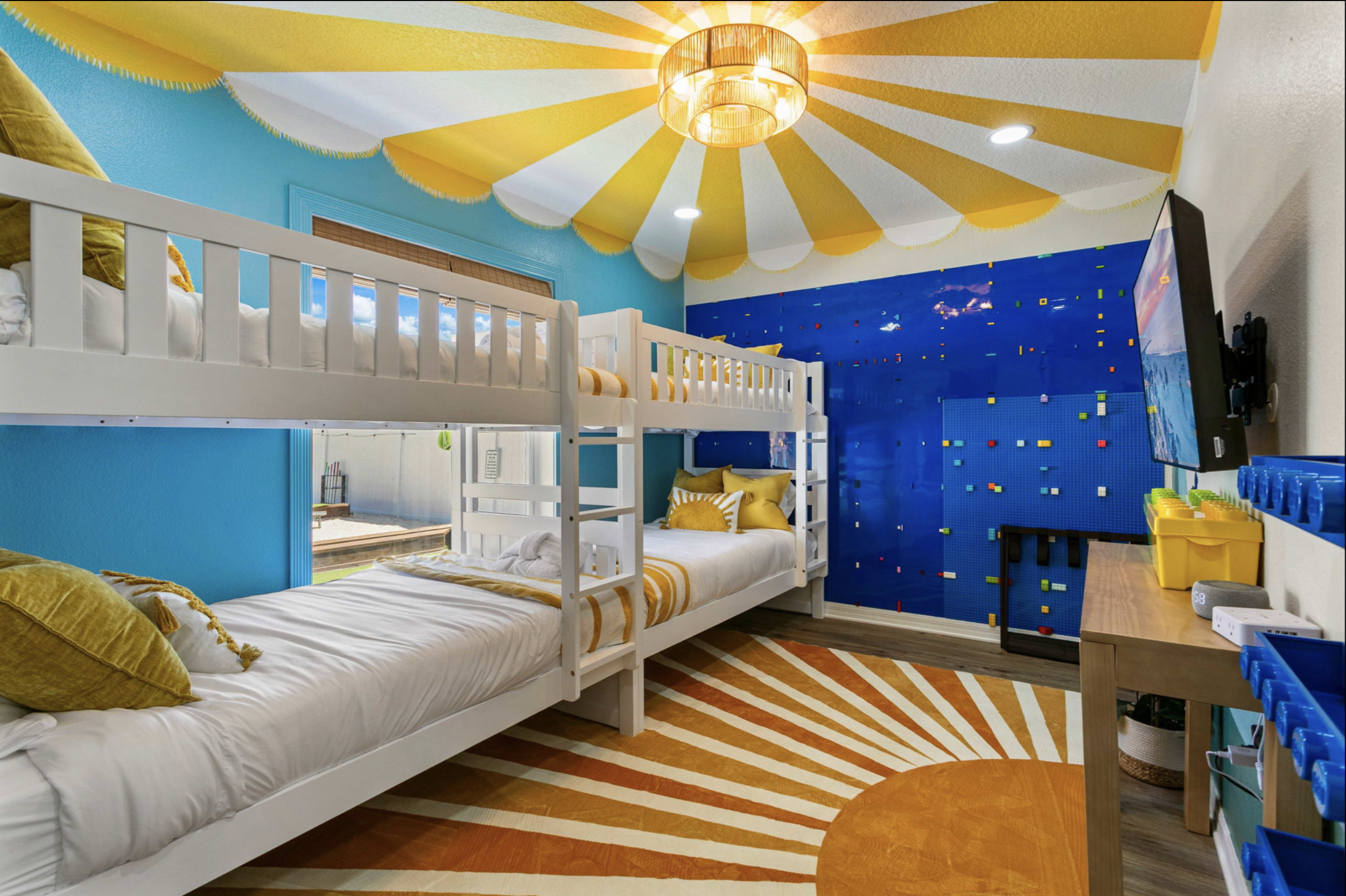 A vibrant children's bedroom with a blue wall decorated with colorful Lego pieces, a ceiling with a yellow and white sunburst pattern, a bunk bed with white frame and yellow pillows, a large window, a pattern carpet with orange and white lines, a wal