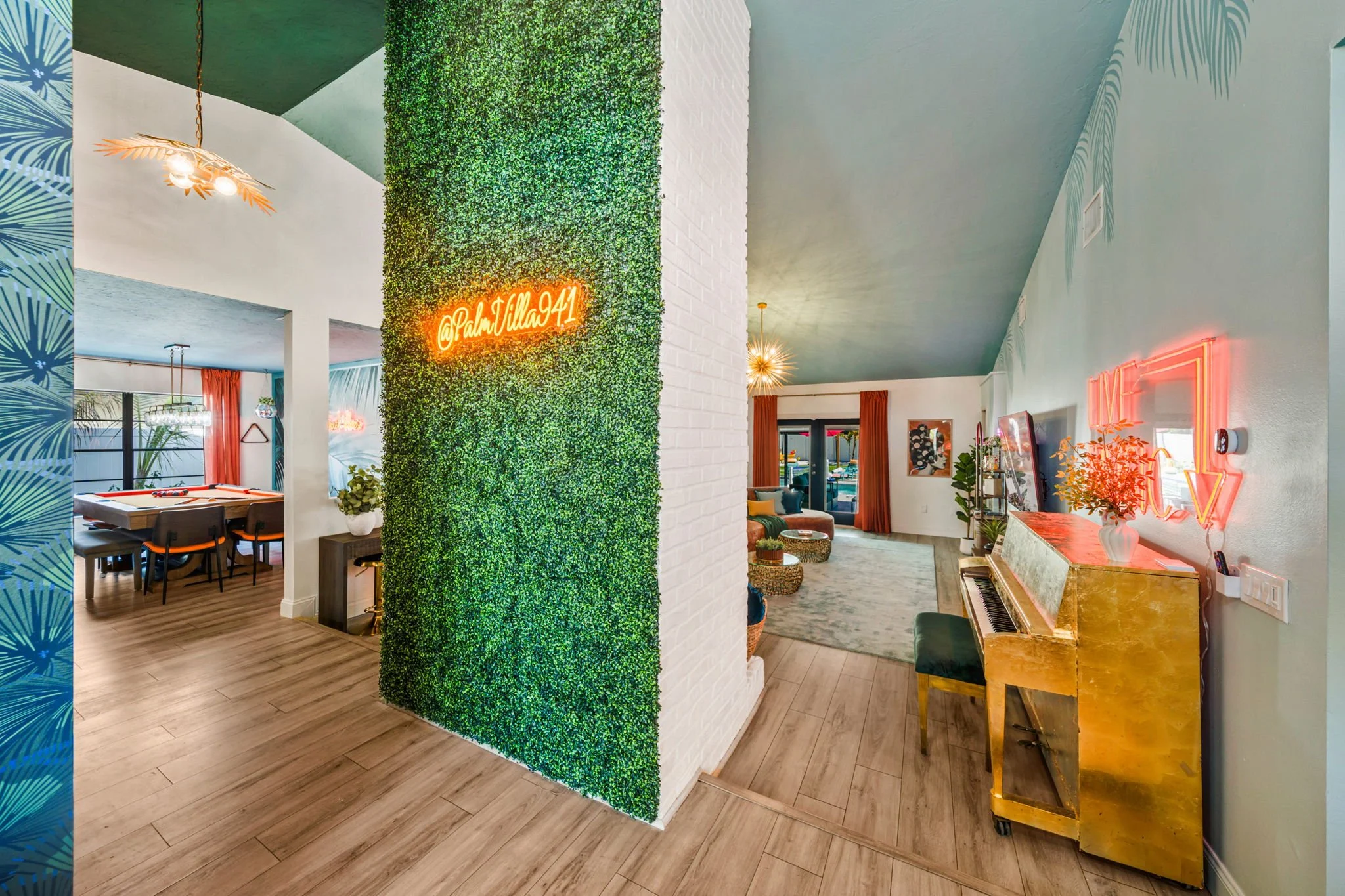 Interior of a colorful living space with a green foliage wall featuring a neon sign reading '@PalmVilla941', a gold piano with a neon 'LOVE' sign on the wall above, and a cozy seating area with rugs, plants, and artwork.