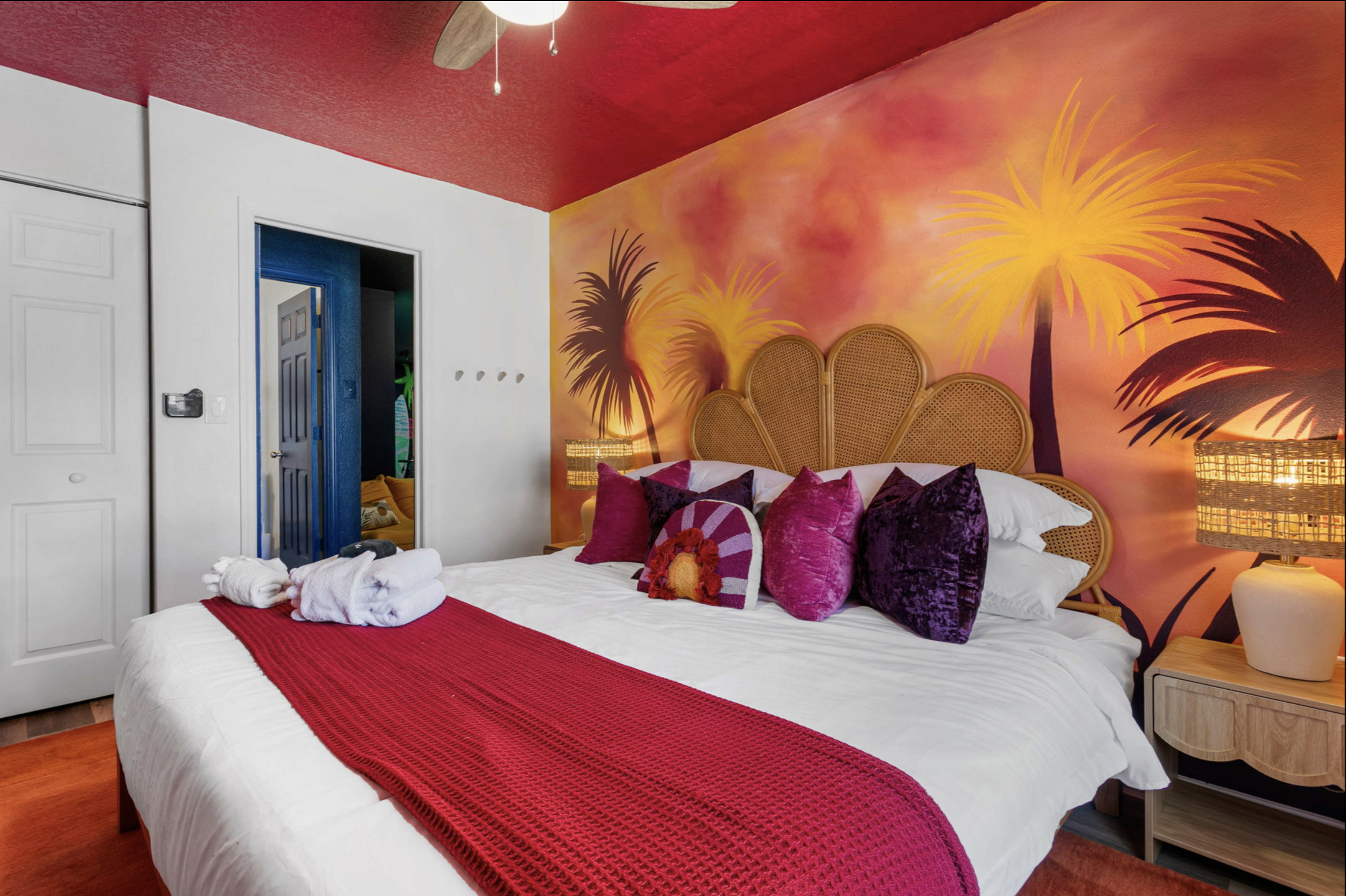 A bedroom with a tropical theme, featuring a wall mural of palm trees and sunset. The bed has a colorful crown-shaped headboard, white sheets, and an assortment of purple and pink pillows. There are lamps on either side of the bed and a red blanket d
