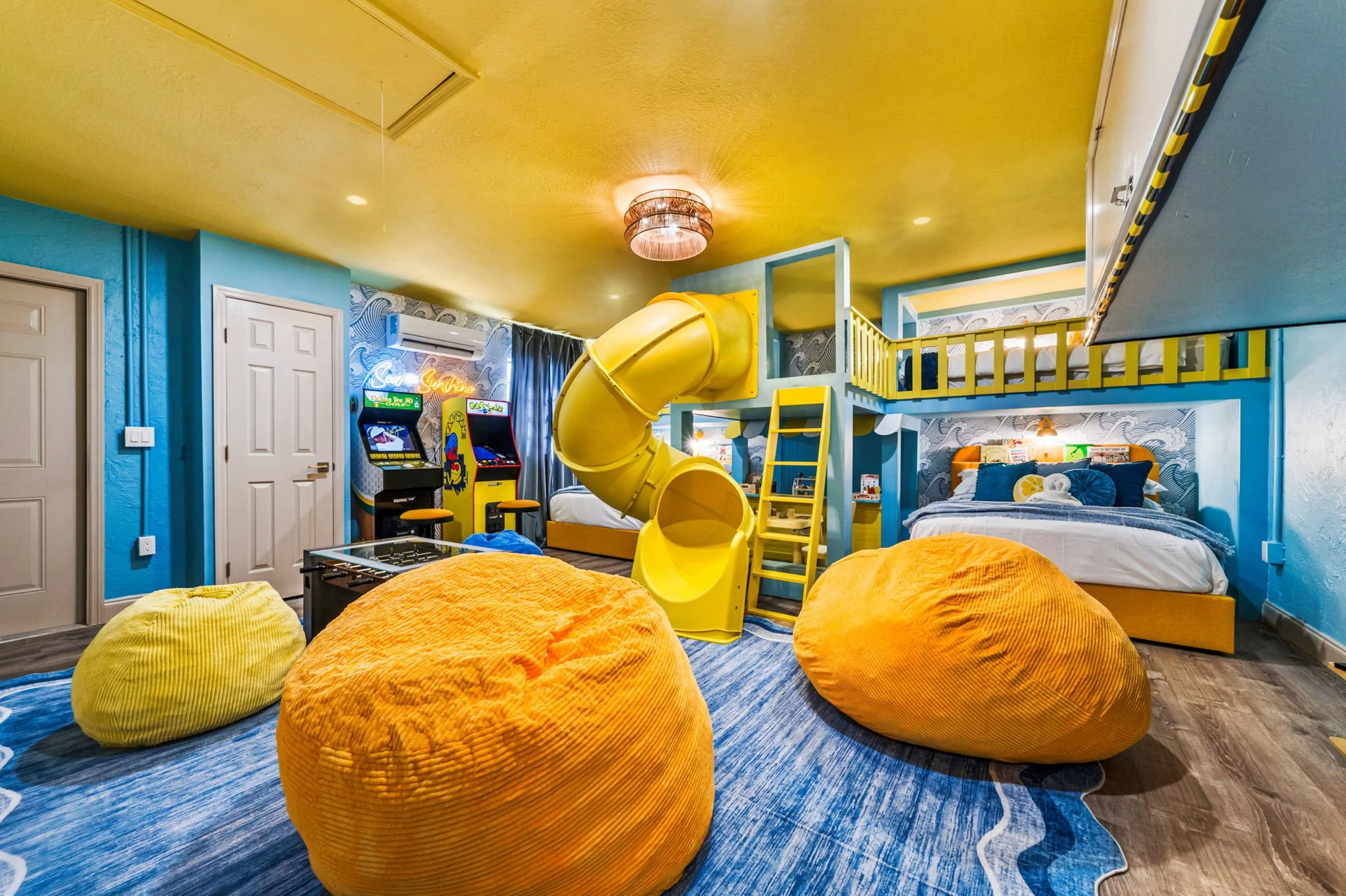 Colorful kids' bedroom with blue walls, yellow ceiling, yellow slide, multiple beds, bean bag chairs, and arcade games.