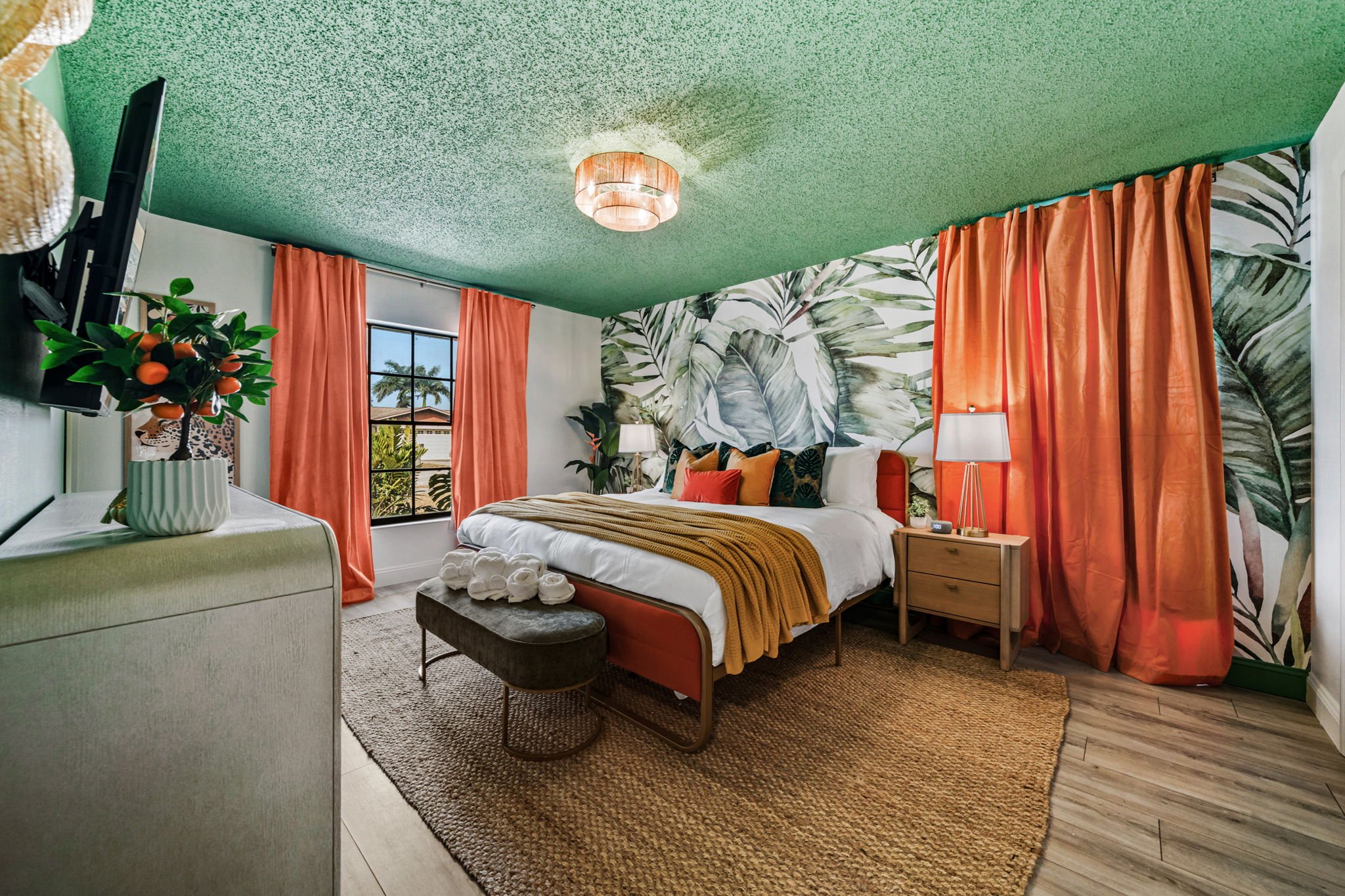 A brightly decorated bedroom with a tropical theme, featuring green textured ceiling, large leaf wallpaper, orange curtains, and a bed with white linens and colorful pillows. There is a small table with a lamp and a window showing palm trees outside.