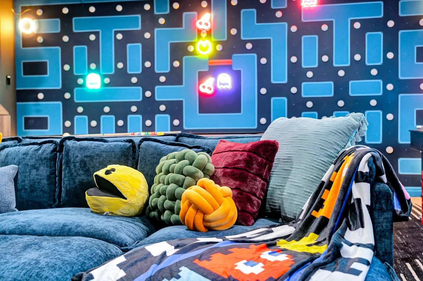 Colorful Pac-Man themed room with a blue velvet couch adorned with pillows, including Pac-Man shaped pillow, and a blanket with Pac-Man pattern. The wall features a large Pac-Man arcade game display with neon lights, and the room has a playful, retro gaming atmosphere.