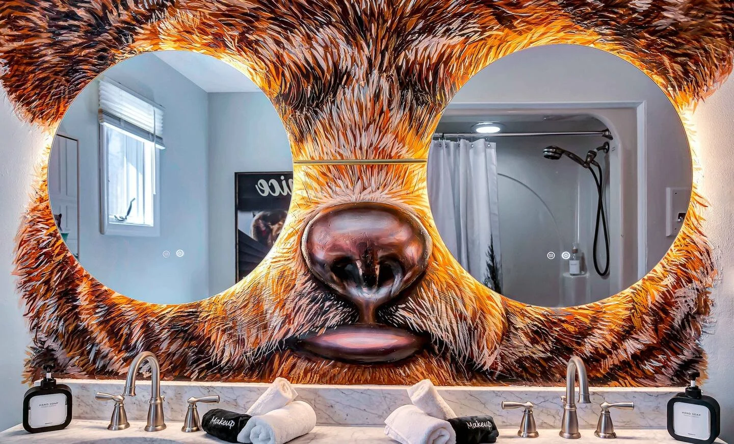 Large mirror on bathroom wall with a bear face design surrounding two circular mirrors, reflecting bathroom interior with a shower, window, and vanity with soap dispensers and towels.