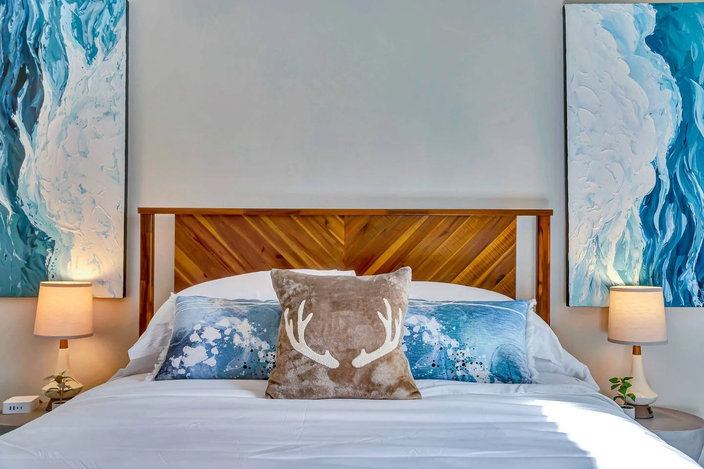 A bedroom with a wooden headboard, two blue and white abstract paintings on the wall, a beige pillow with white antlers, and two bedside lamps with small plants.