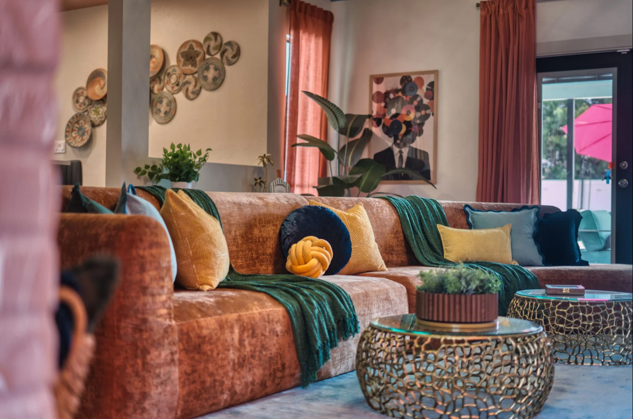 Living room with a brown velvet sectional sofa, colorful pillows, green throws, decorative coffee tables, houseplants, wall art, and a door leading to an outdoor patio with a pink umbrella.